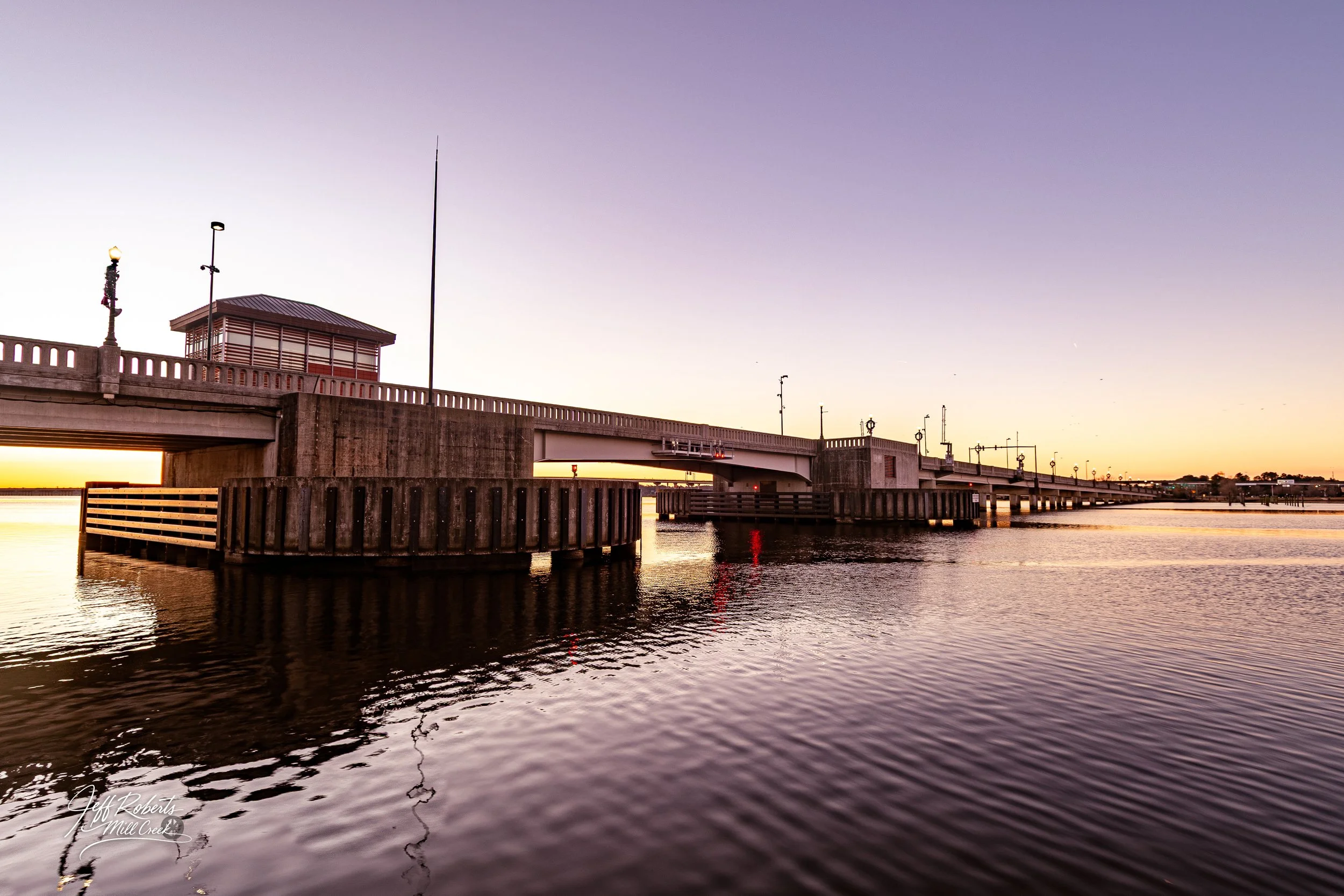 A bridge over a body of water during sunset with a small control building and decorative streetlights on it.