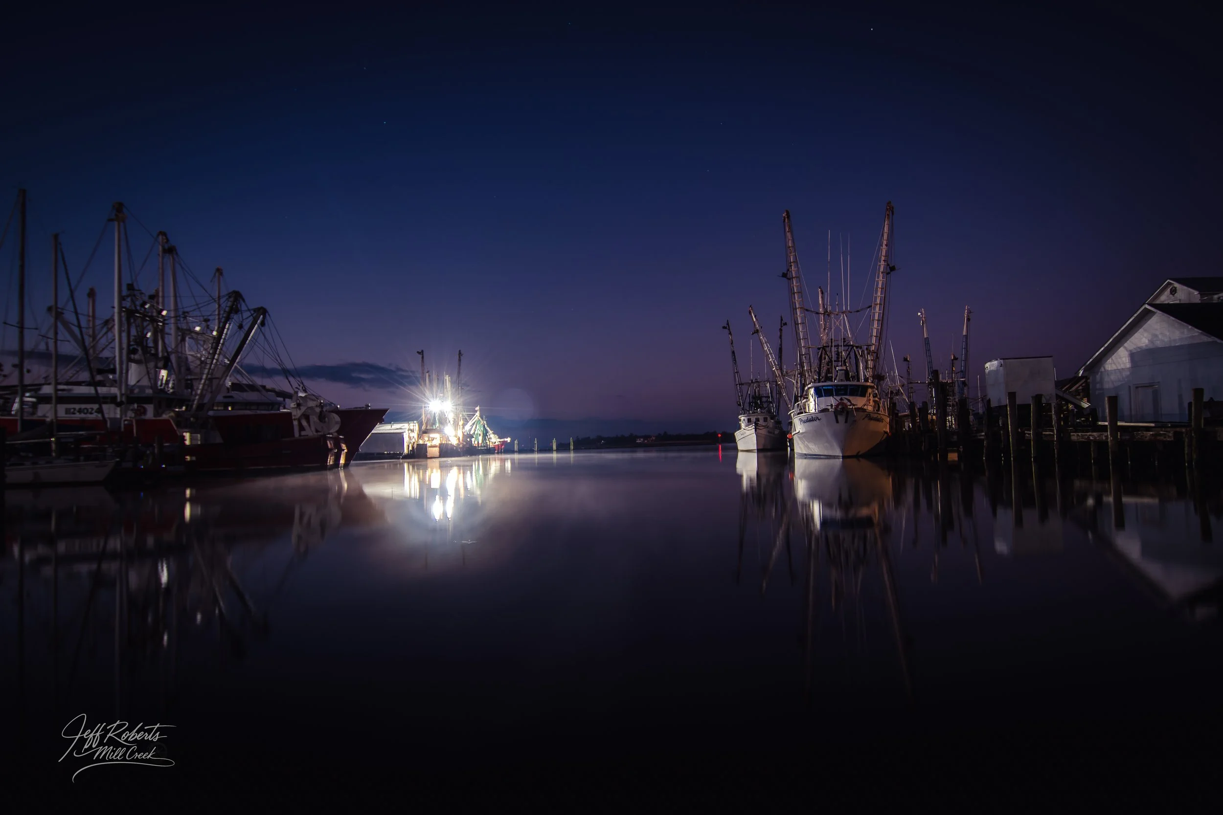 Boats docked at a harbor during nighttime, with calm water reflecting the boats and a deep blue sky with stars.