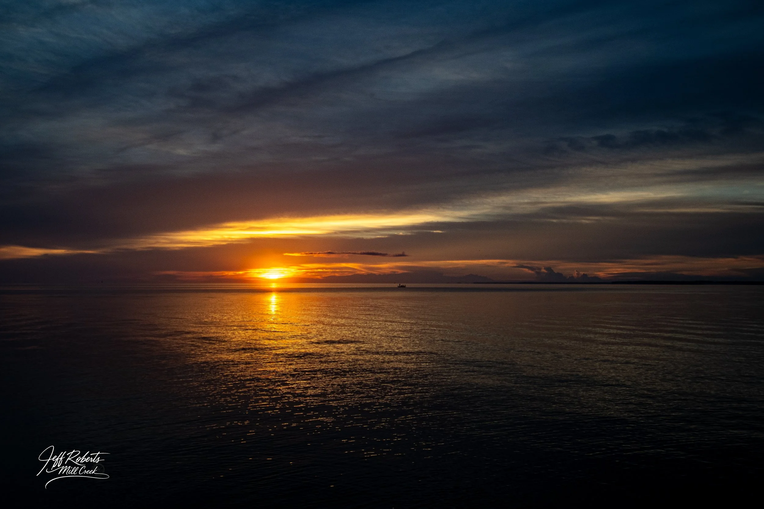 Sunset over a calm body of water with a cloudy sky, a small boat silhouette on the horizon, and the sun partially obscured by clouds.