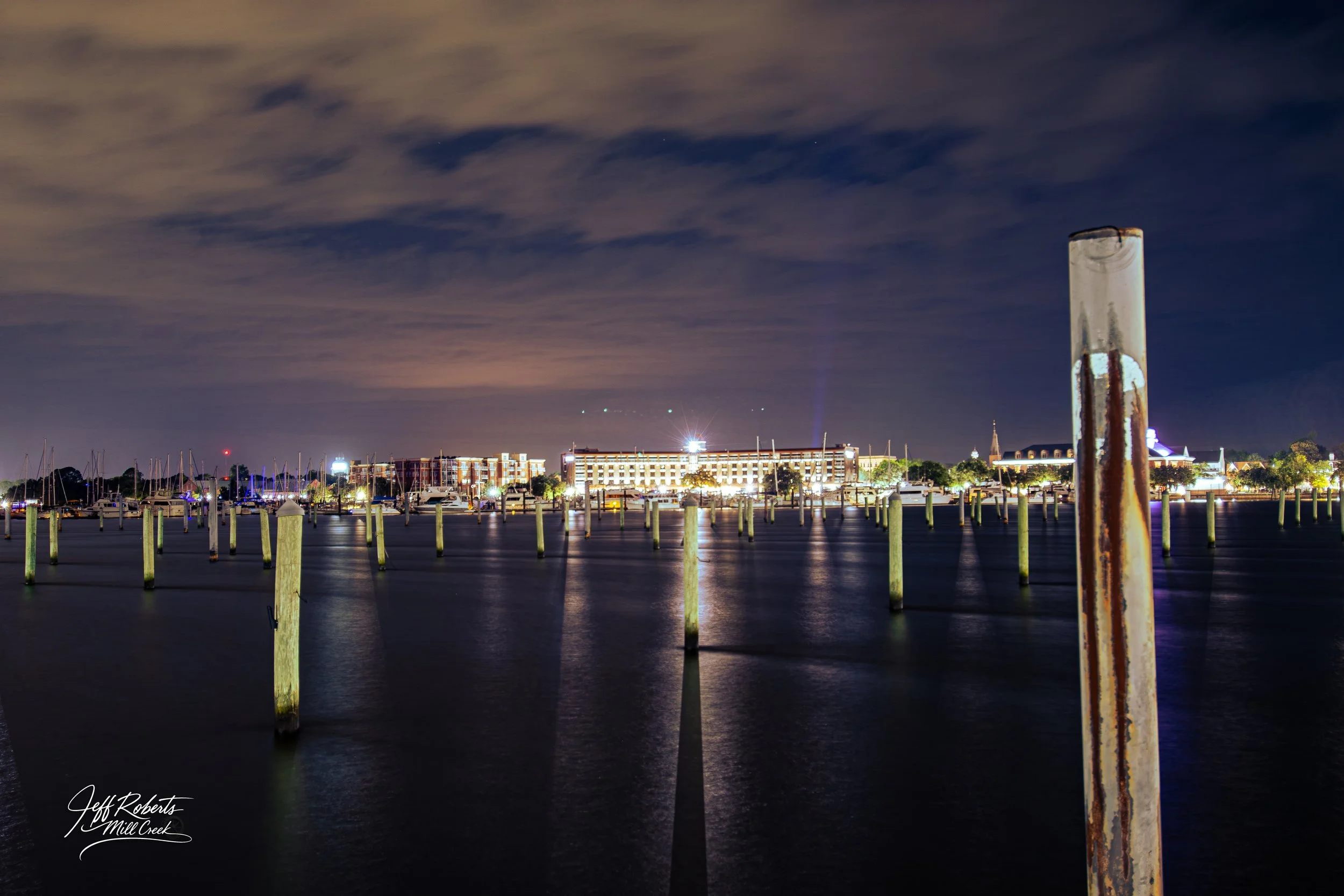 Nighttime view of a marina with boats, pilings, and illuminated buildings in the background.