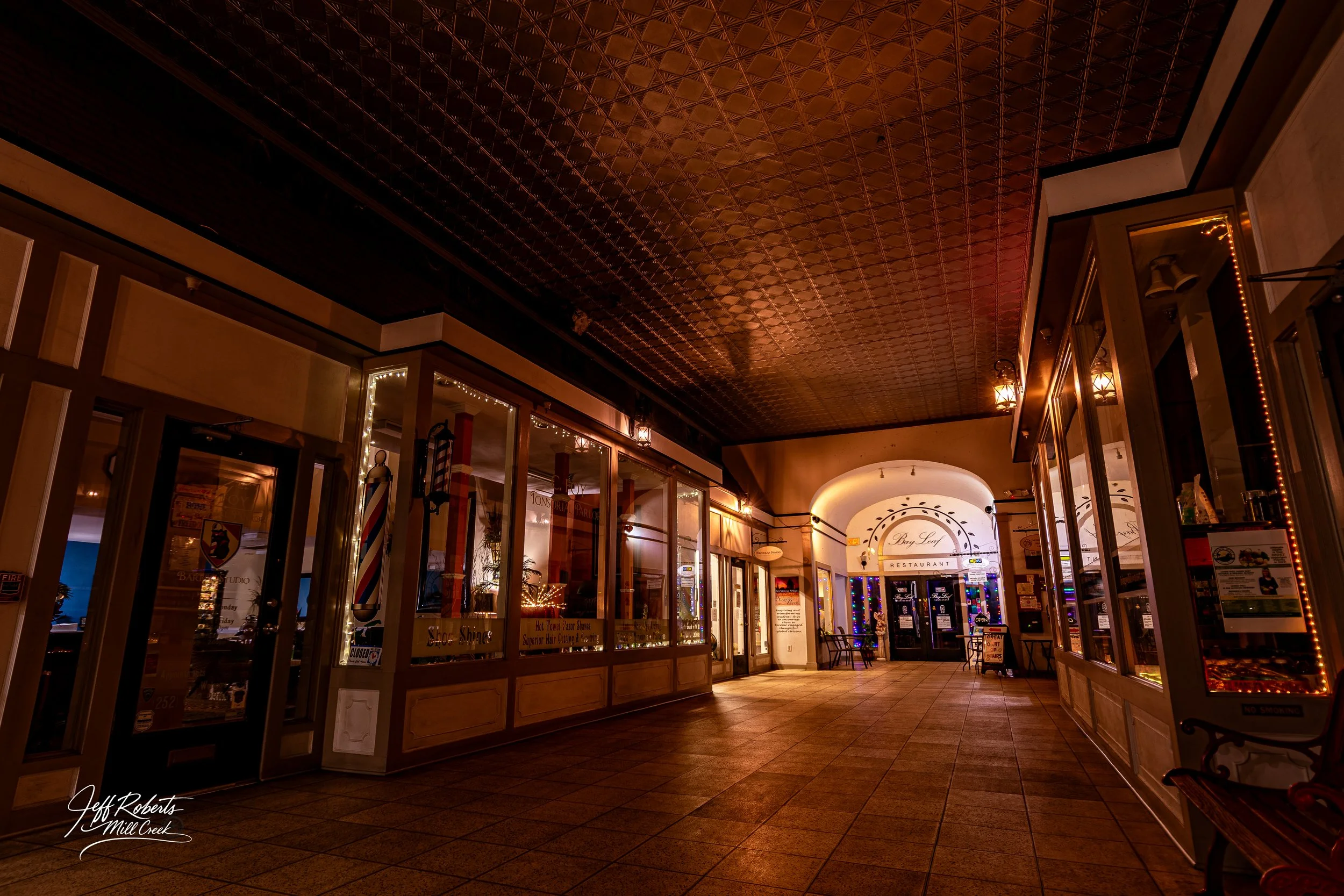 Empty shopping corridor with closed storefronts, warm lighting, and decorative ceiling, leading to a restaurant entrance in distance.