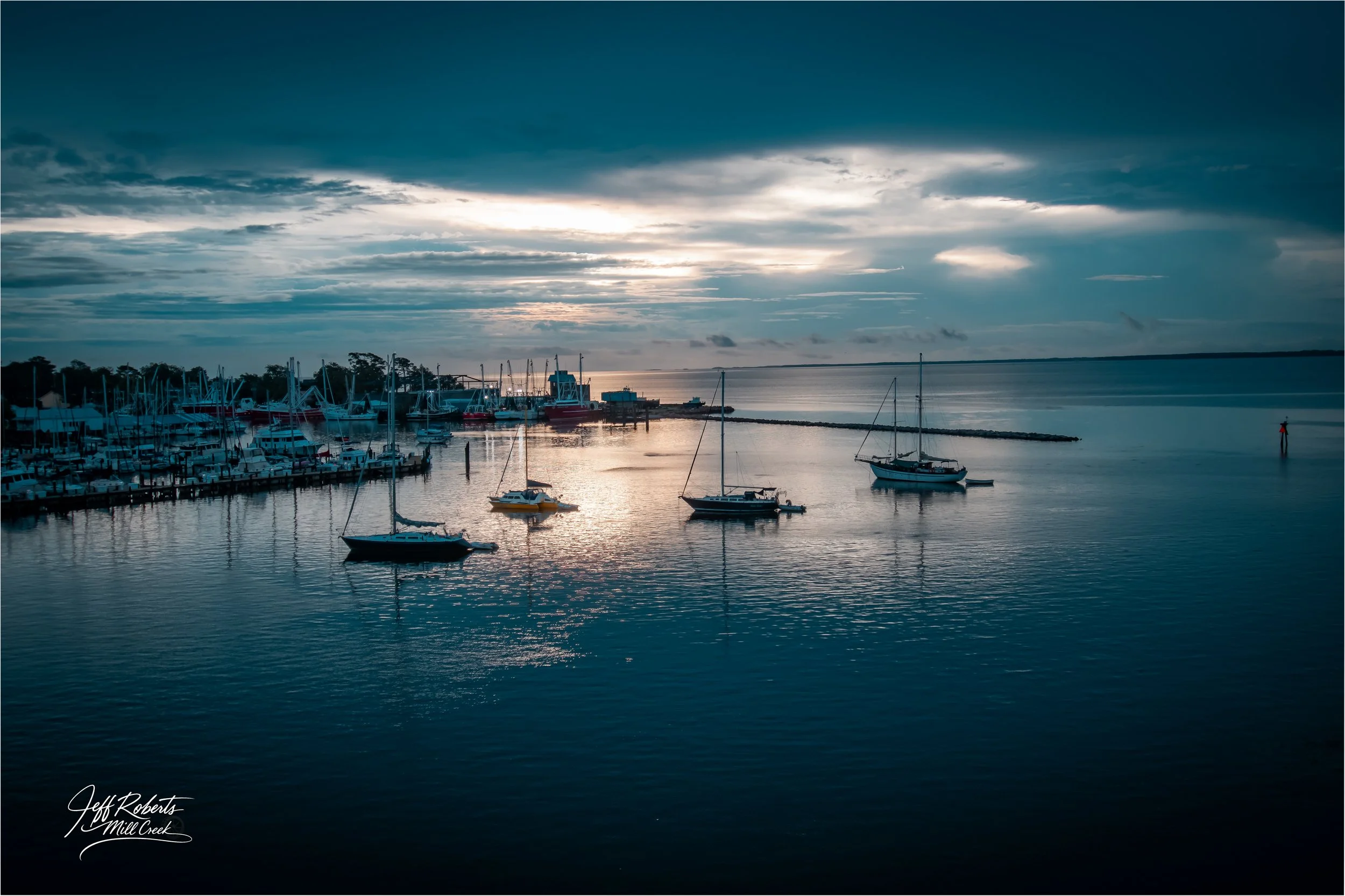 A serene harbor scene at sunset with several sailboats and yachts anchored on calm water, a cloudy sky with some sunlight peeking through, and a marina in the background.