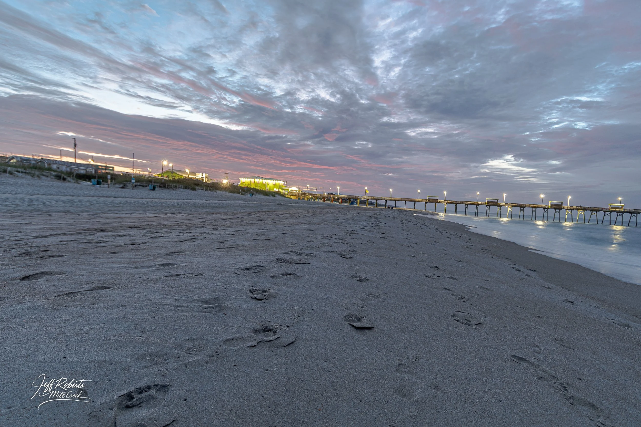 Footprints on a sandy beach at sunset near a pier with lights and buildings in the background.