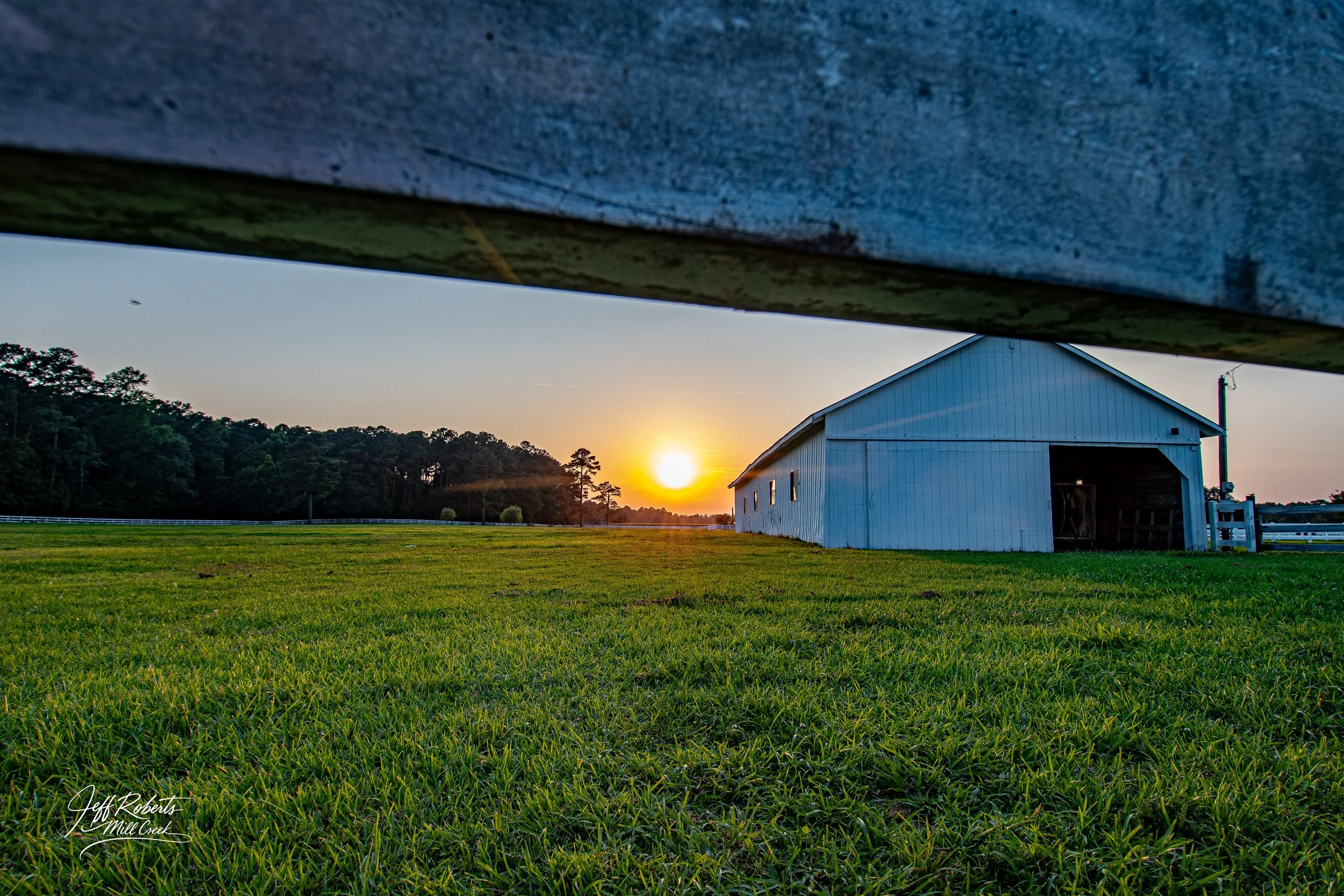 Sunset over a grassy field with a white barn to the right and a blue fence in the foreground.