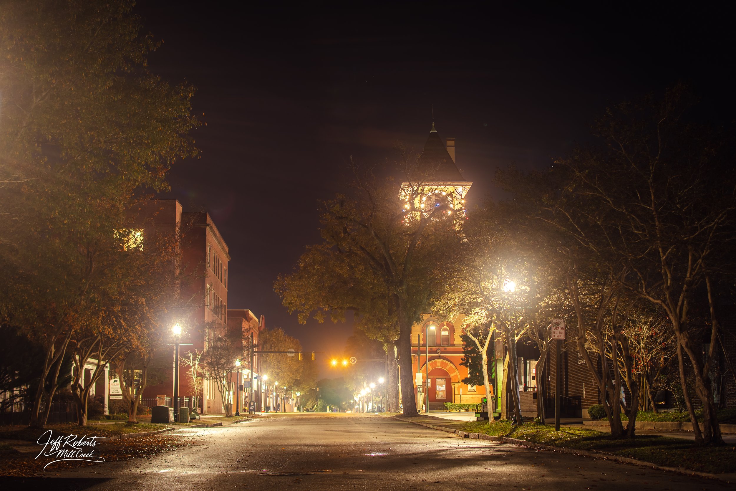 A quiet, empty street at night with streetlights illuminating the sidewalk and trees. A clock tower building is visible in the background, lit up against the dark sky. The scene has a peaceful, small-town feel.