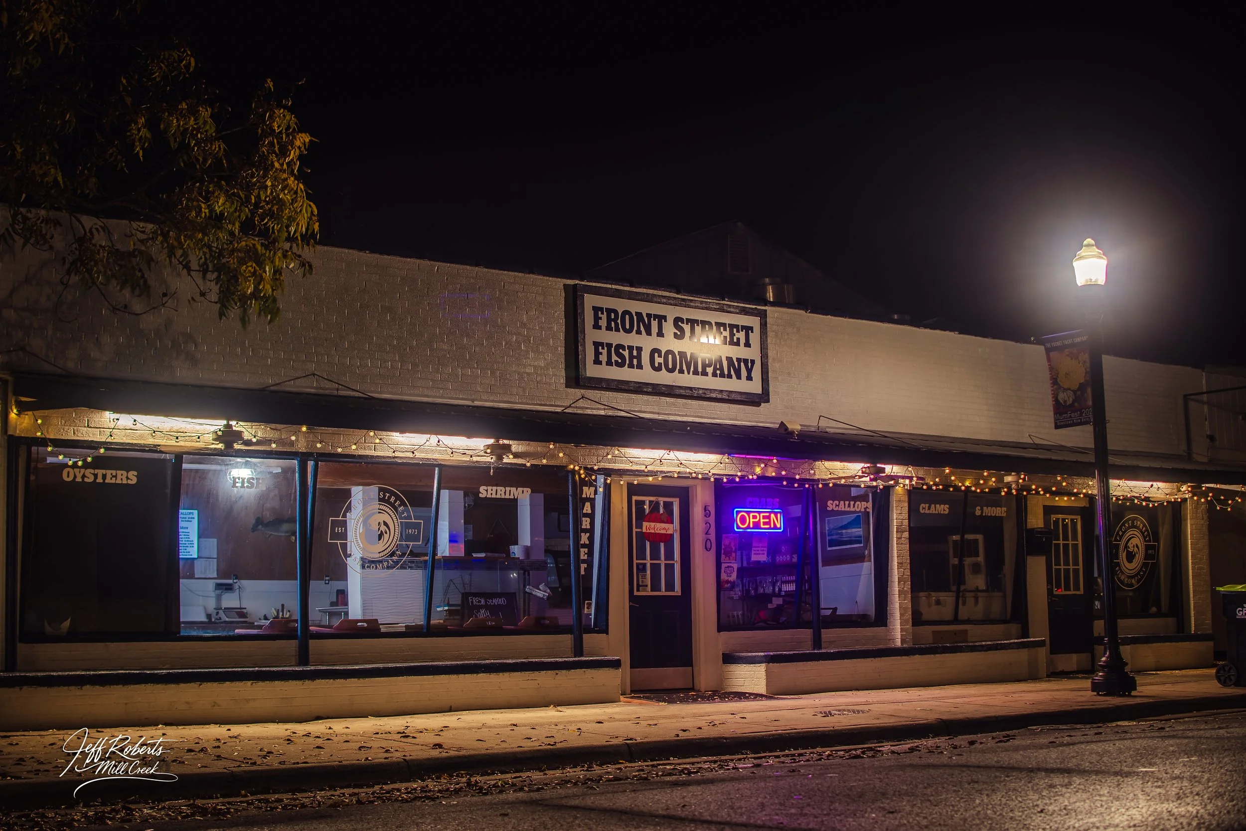 Night view of a seafood restaurant called Front Street Fish Company with an illuminated 'OPEN' sign, string lights, and a clear window showcasing the interior.