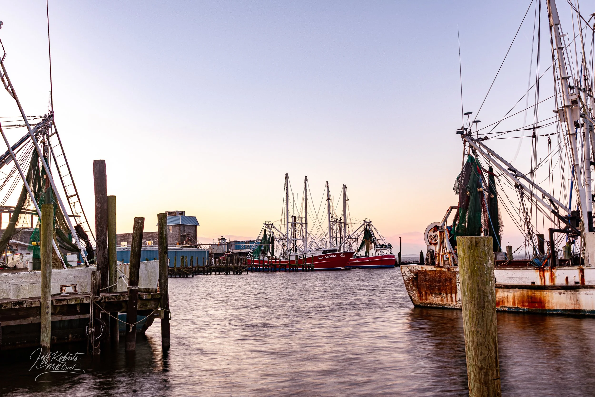 A harbor scene at dusk with several fishing boats docked, some with rust and green covers, wooden posts, and a small building in the background.