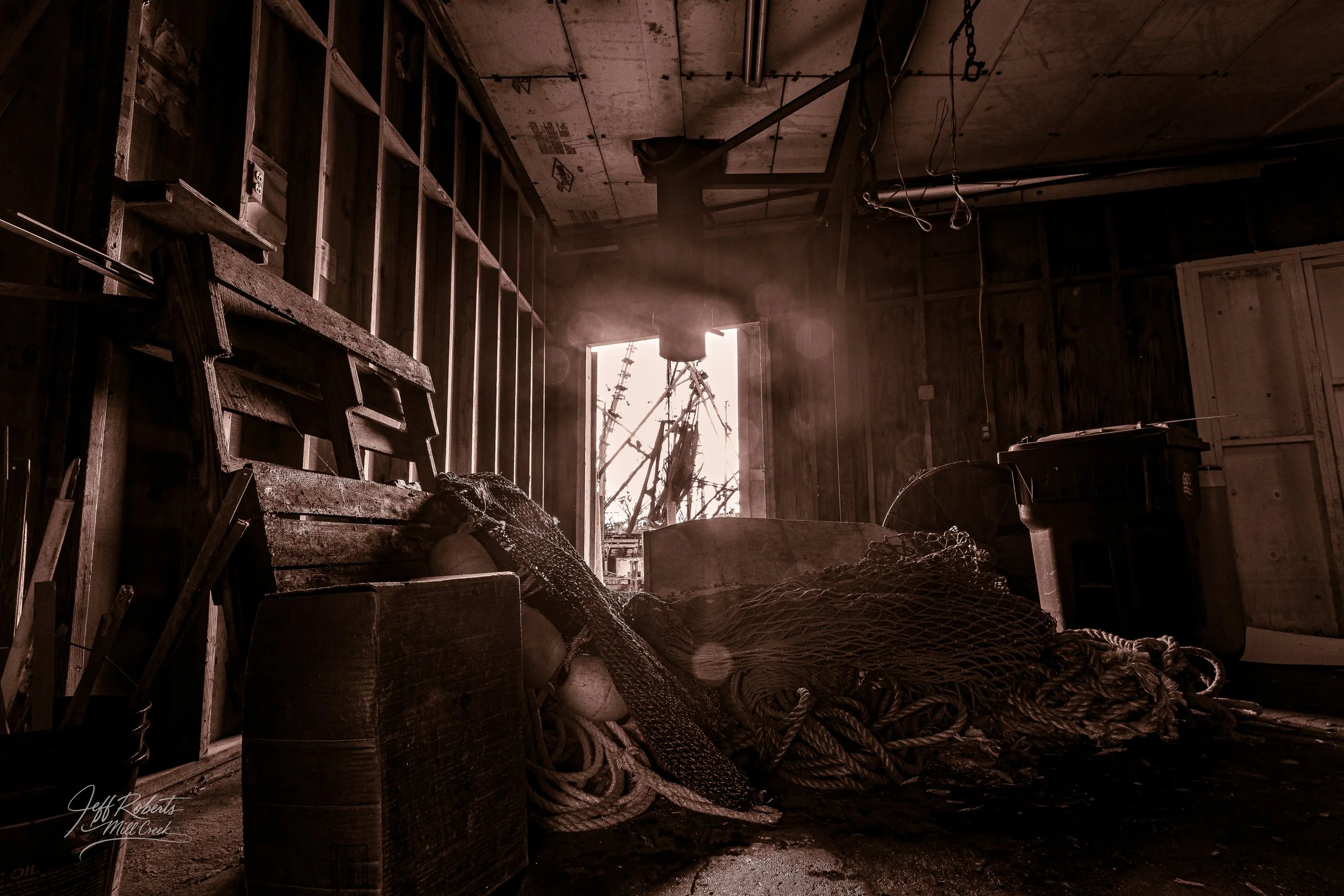 Interior of a rustic wooden shed with fishing nets, a wooden chair, and a window showing damaged utility poles outside.