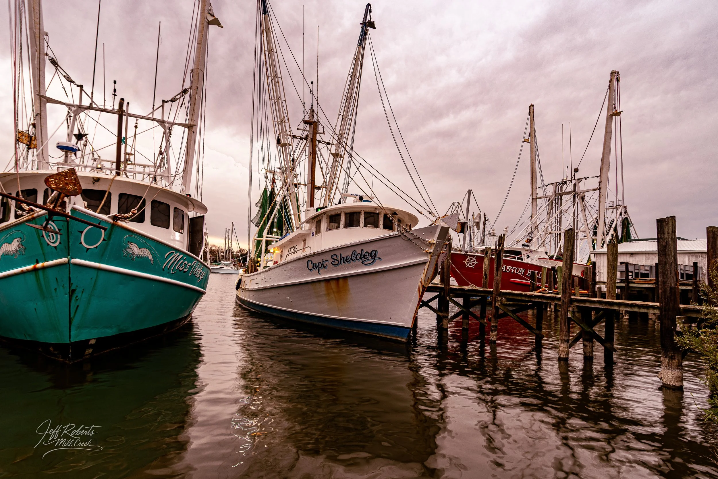 Several boats docked at a pier on a cloudy day, with reflections visible in the water.
