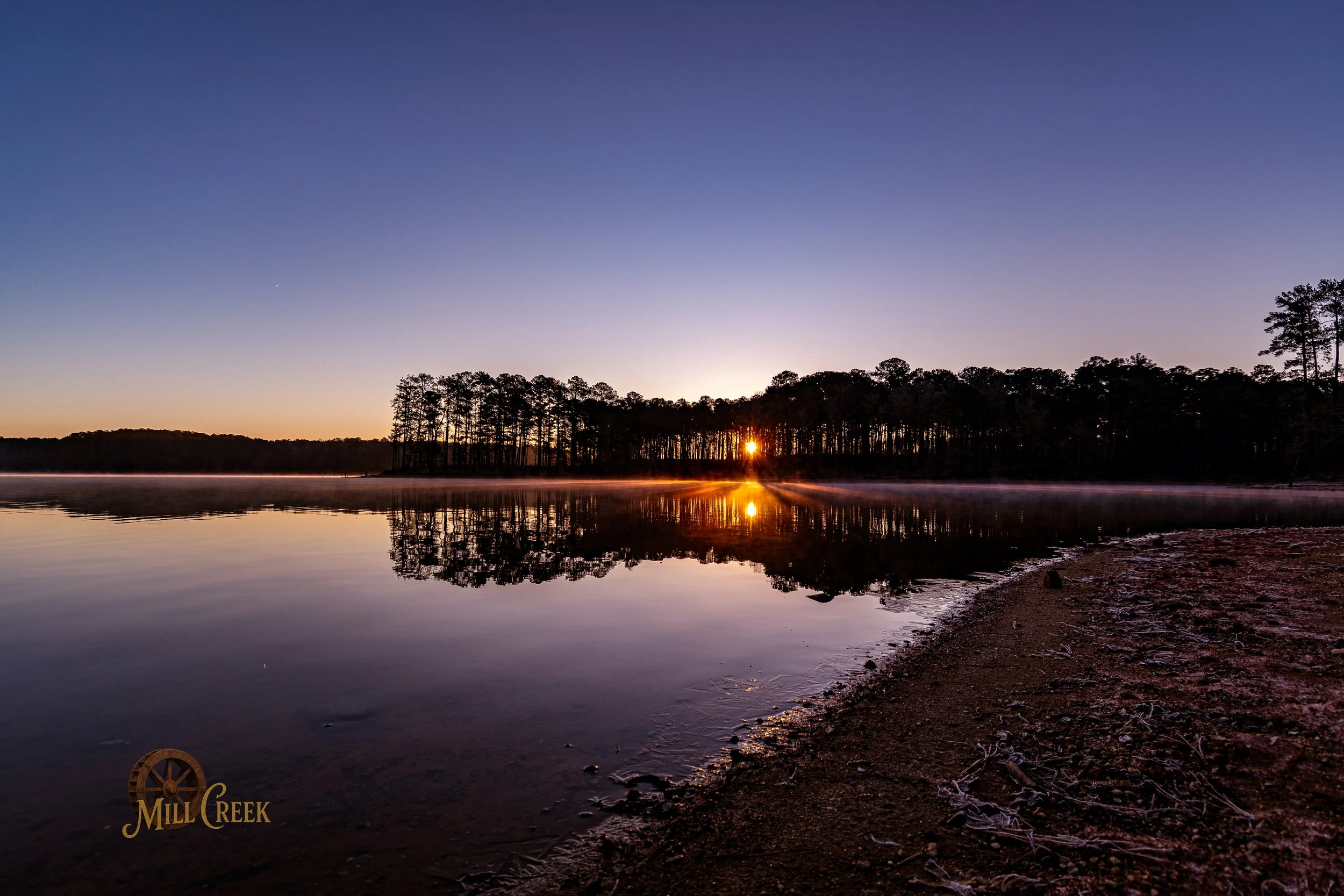 Sunset over a calm lake with a tree-lined horizon, reflections on the water, and a clear sky with a faint celestial body visible.