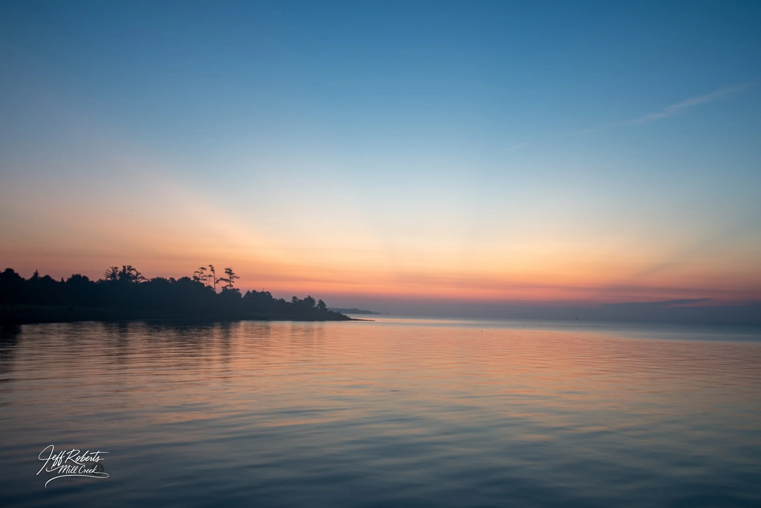 Serene sunset over a calm river with reflections of the sky on the water, silhouette of trees on the horizon.