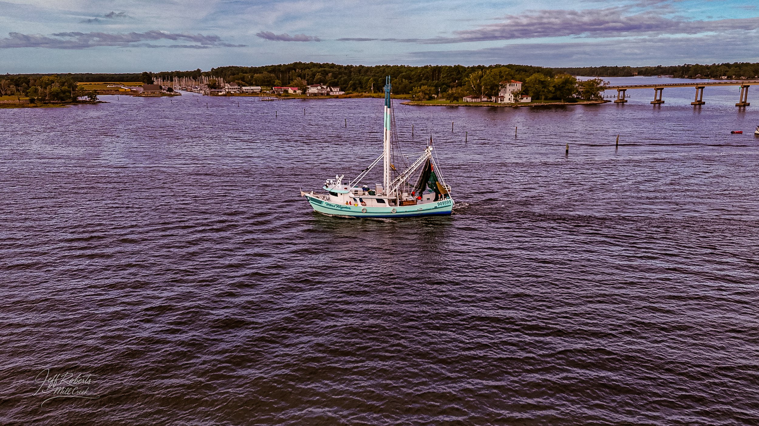 A fishing boat floating on a calm river with small waves. In the background, there are houses, trees, and a bridge crossing the river under a cloudy sky.