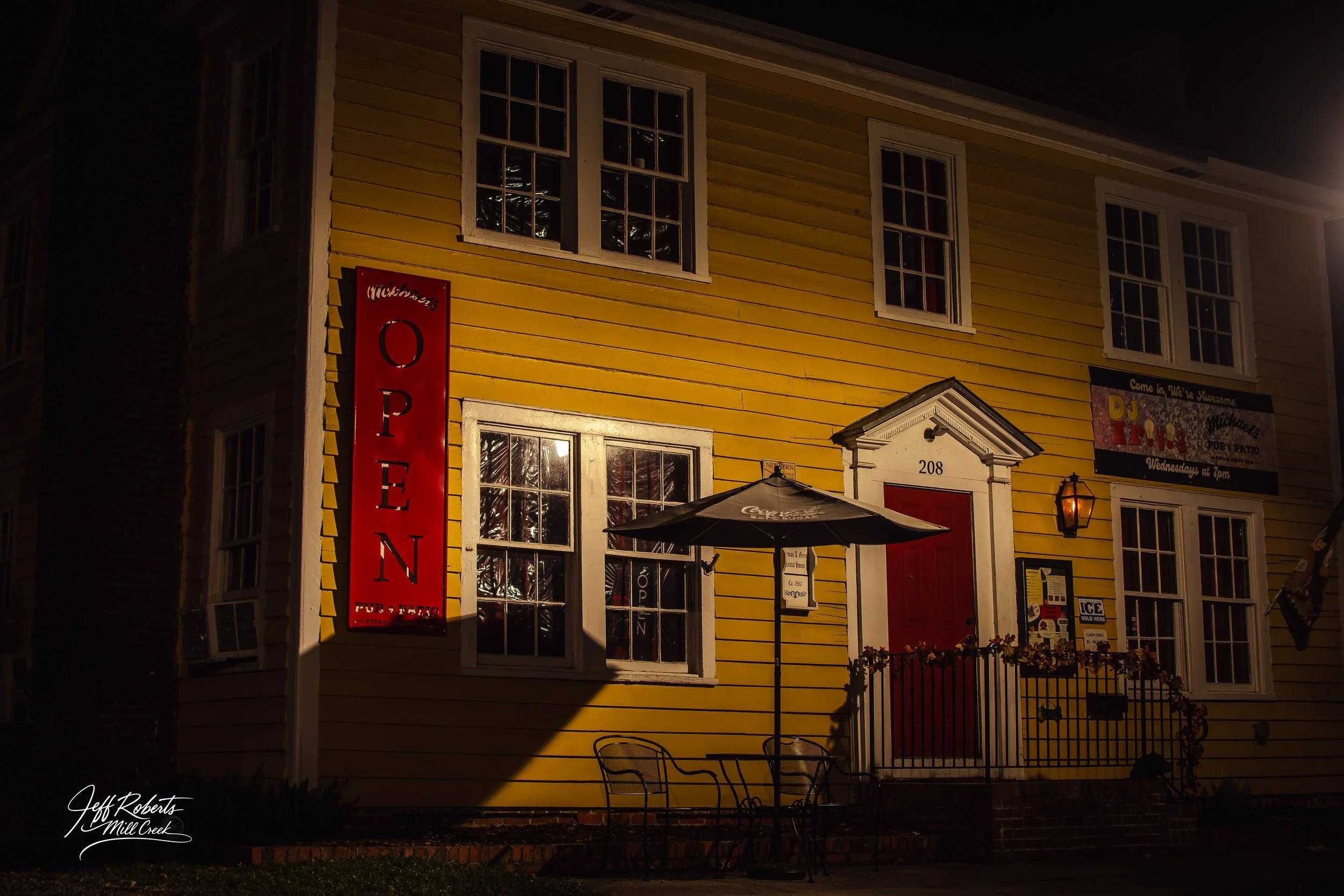 Night view of a bright yellow building with white trim and red doors, featuring a large red 'OPEN' sign, outdoor seating with a black umbrella, and signs indicating it is a pub and patio with entertainment, along with a wall-mounted lantern lighting 