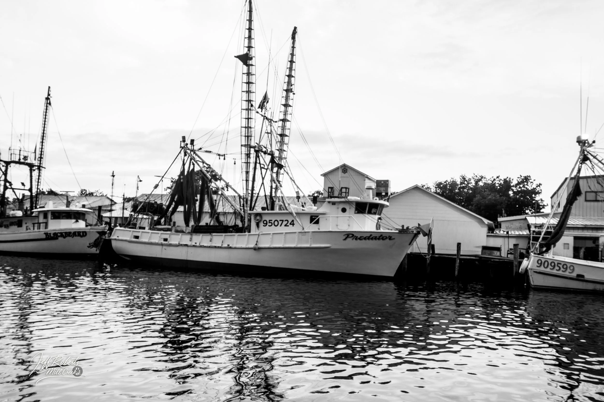 Black and white photo of boats docked at a harbor, with a house and trees in the background.