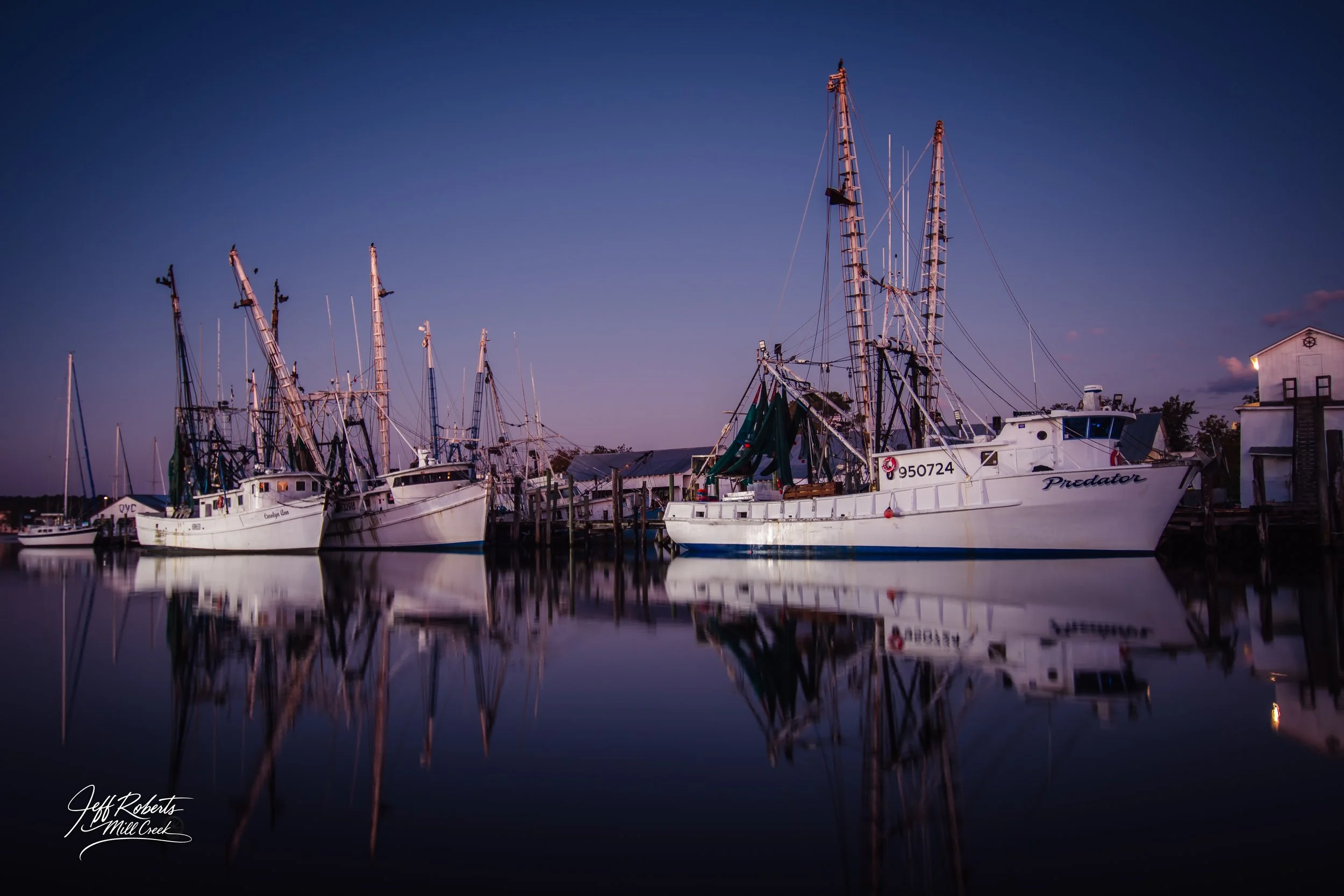 Several boats docked at a marina during twilight with calm water reflecting the boats and sky.