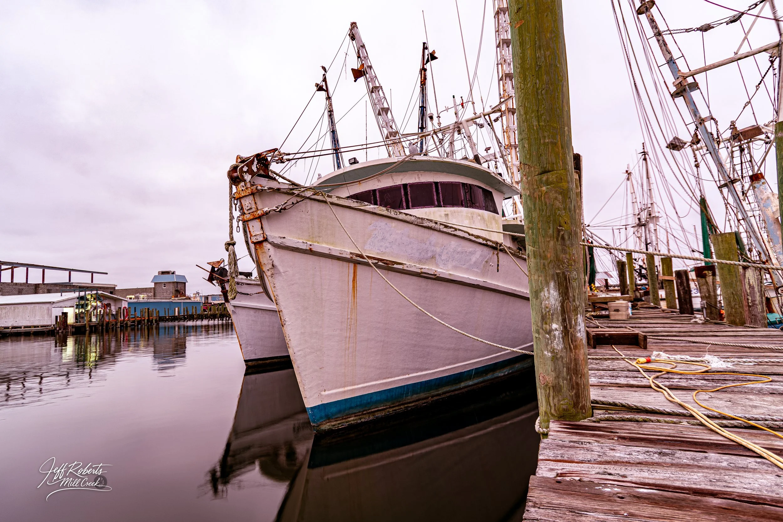 An old, weathered white boat docked at a wooden pier, with rusty chains and ropes, in a harbor with water reflecting the boat and a cloudy sky overhead.