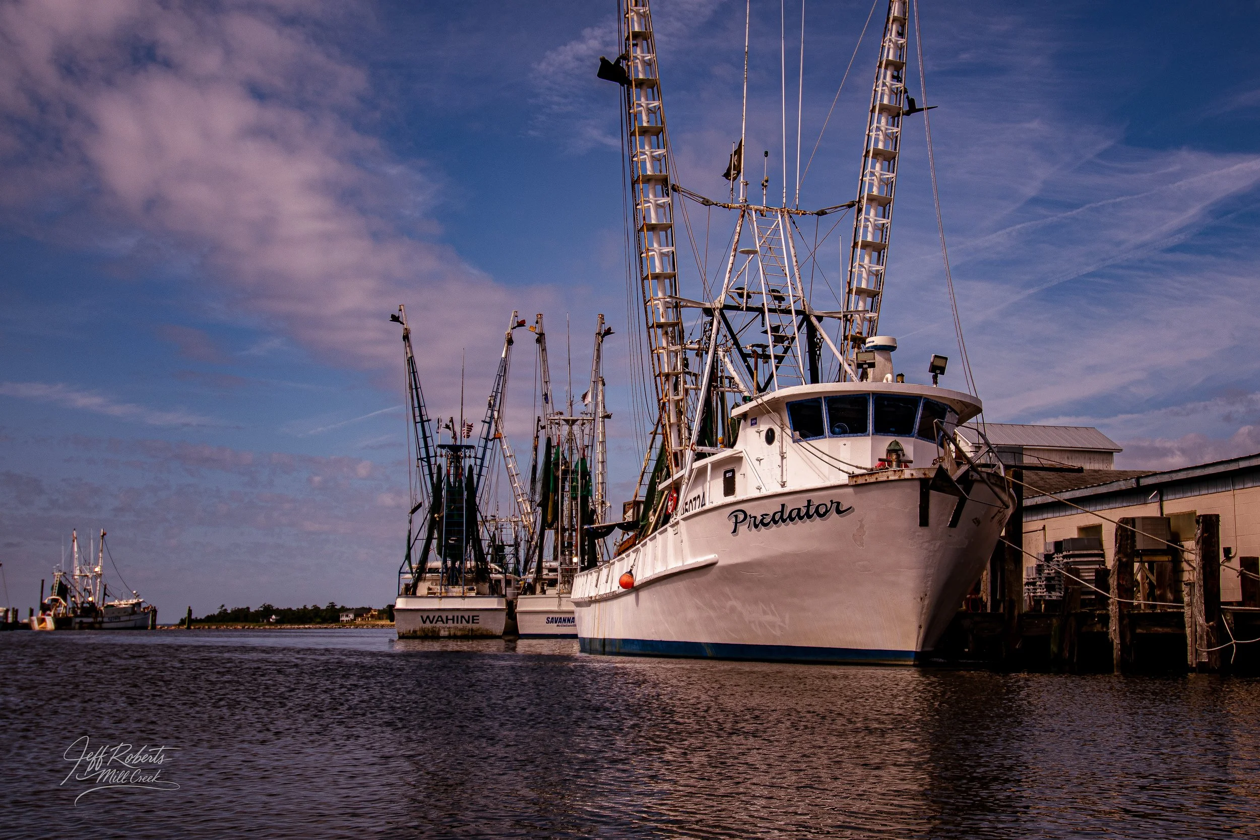 Several fishing boats docked at a harbor with calm water and a partly cloudy sky in the background.