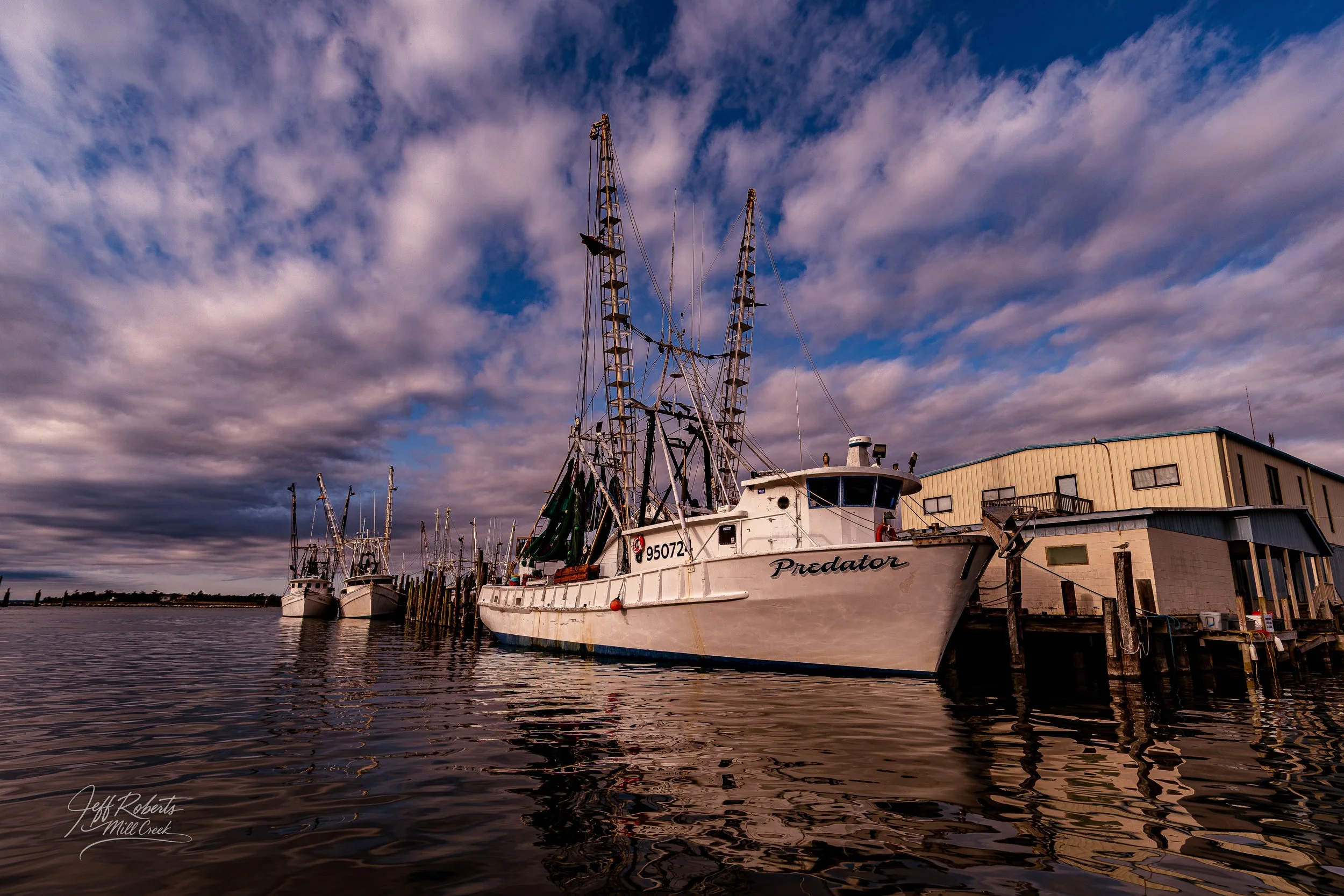 A white fishing boat named Predator docked at a pier with other boats in the background, under a cloudy sky during sunset.