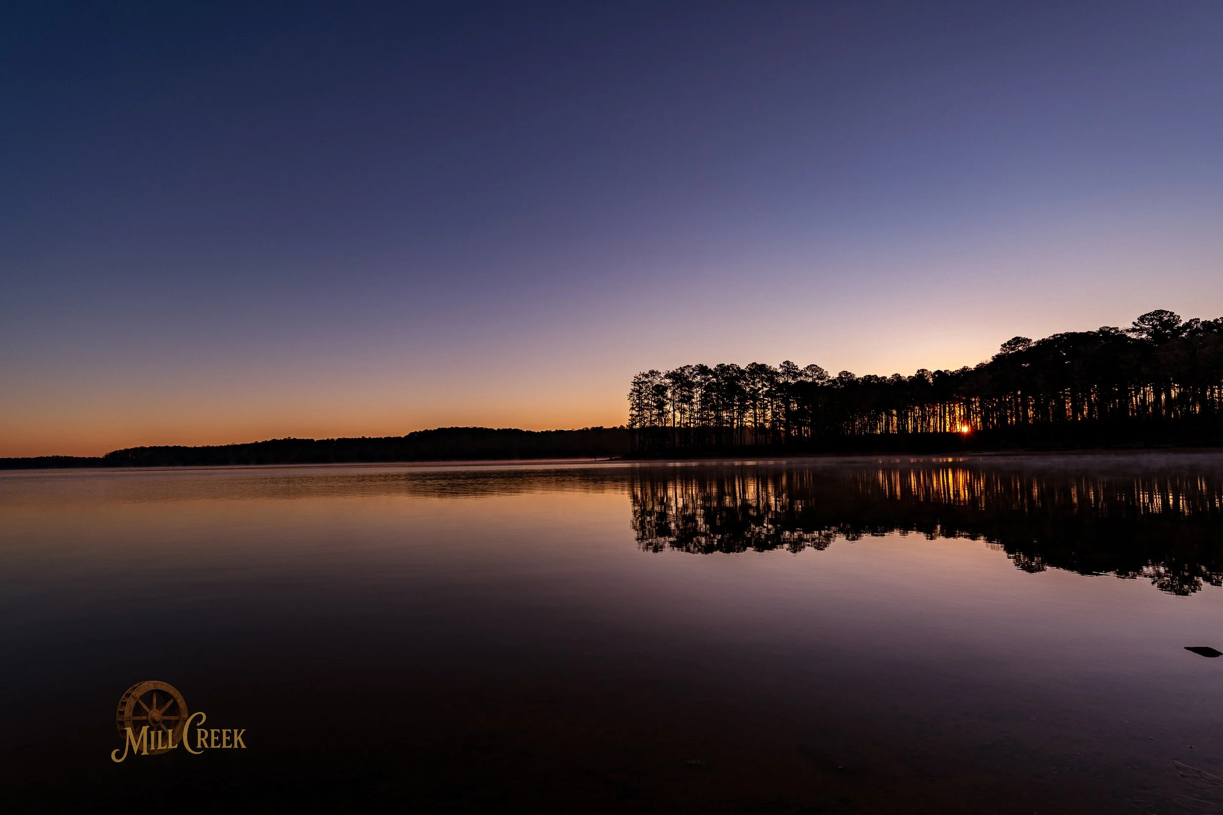 A serene lake at sunrise with a silhouette of trees on the horizon and their reflection on the calm water. The sky transitions from dark blue to soft orange. The Mill Creek logo is in the bottom left corner.