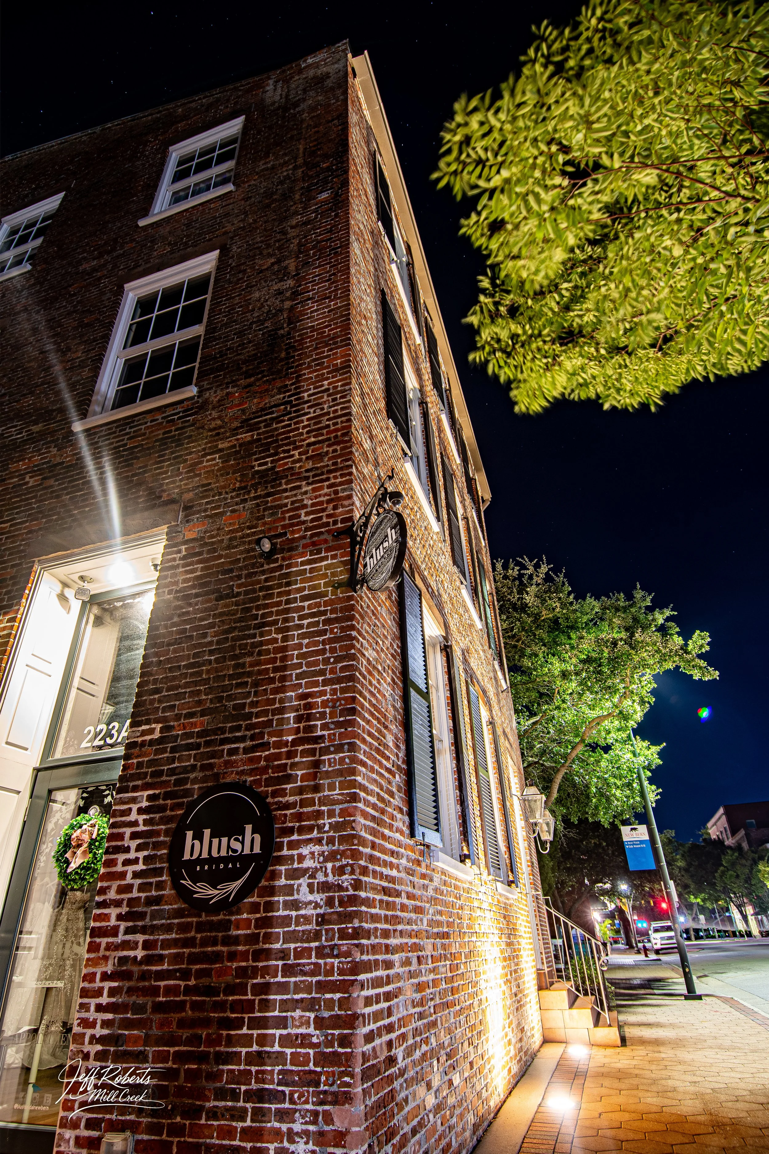 Nighttime exterior view of a brick building with a sign for 'blush bridal' on the wall, illuminated by exterior lighting, with trees and street visible in the background.