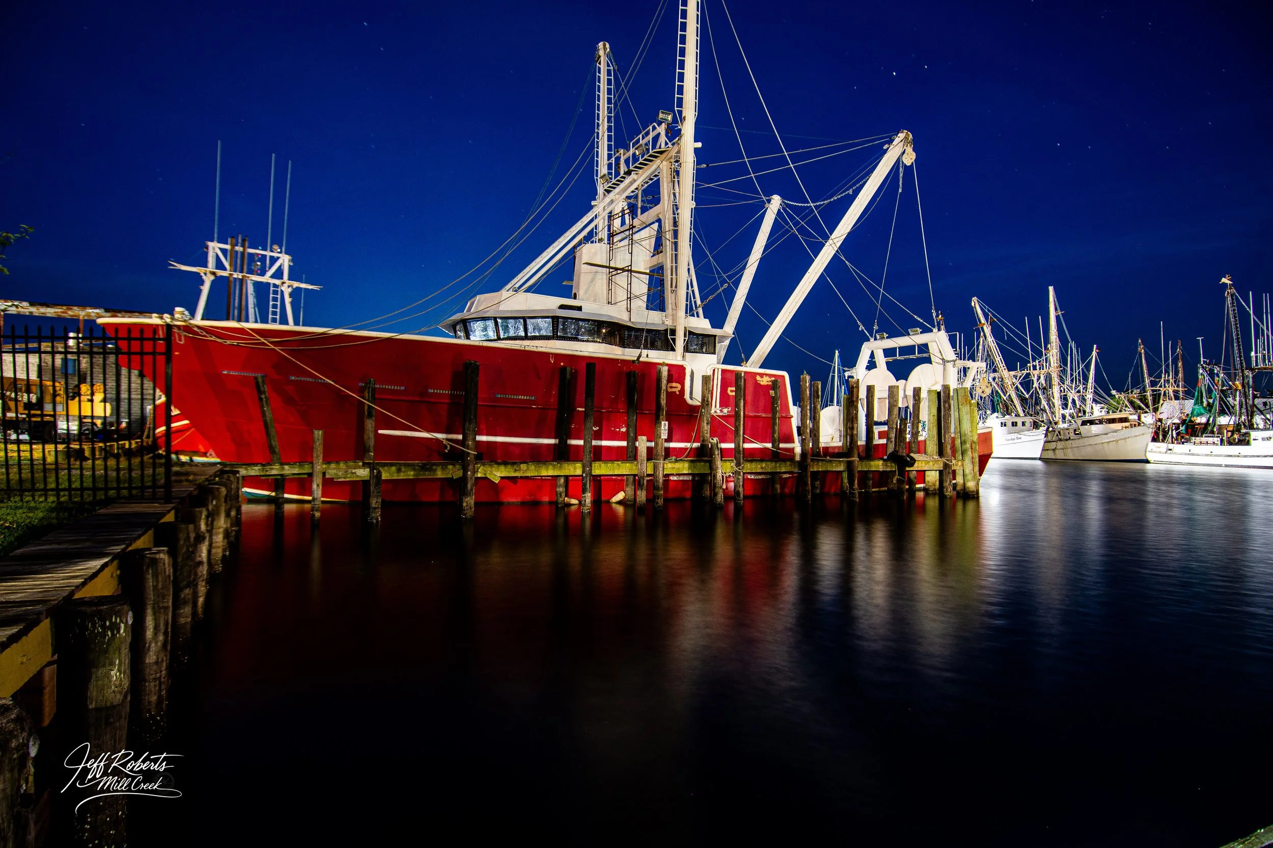Nighttime view of boats docked at a marina, with a prominent red fishing boat in the foreground and several other white sailboats in the background, calm water reflecting the boats, and a dark starry sky above.