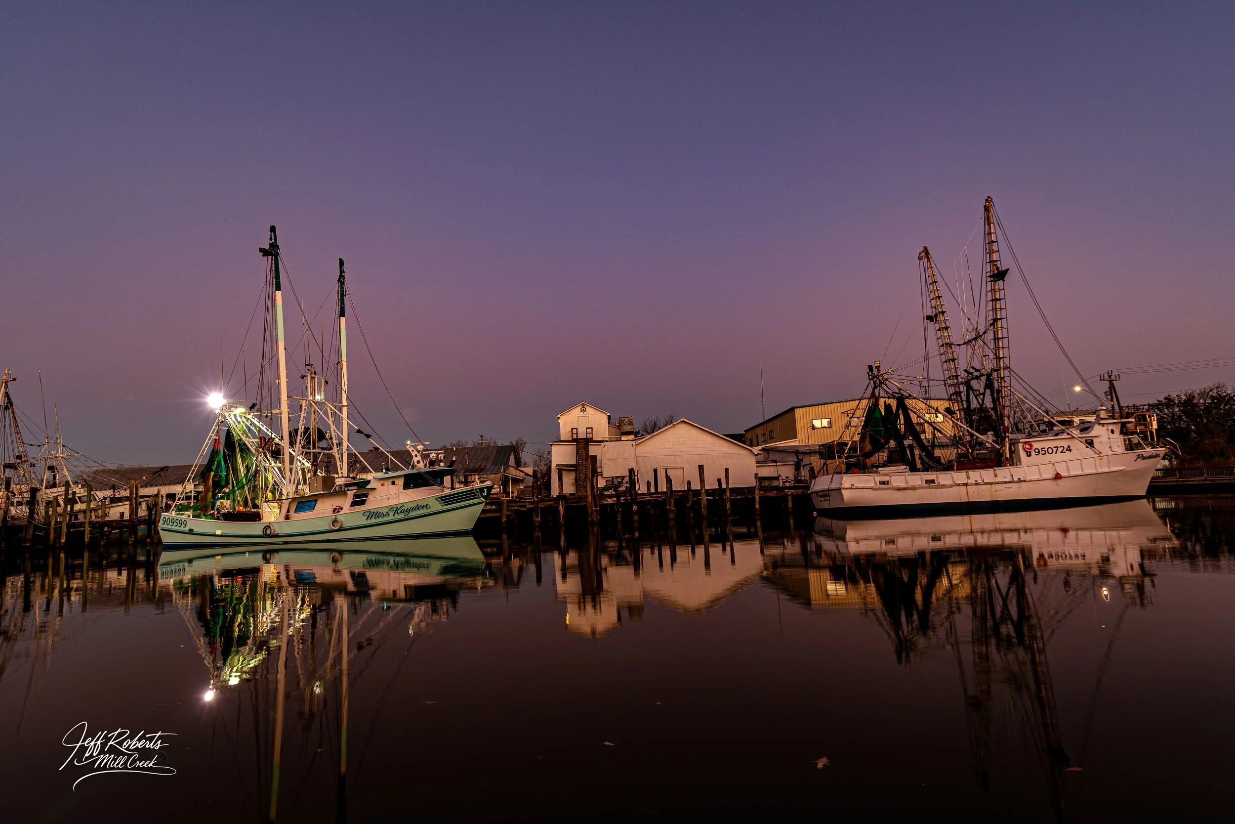 Two boats docked at a marina during dusk with calm water reflecting the boats and structures along the pier, including small buildings and industrial equipment.