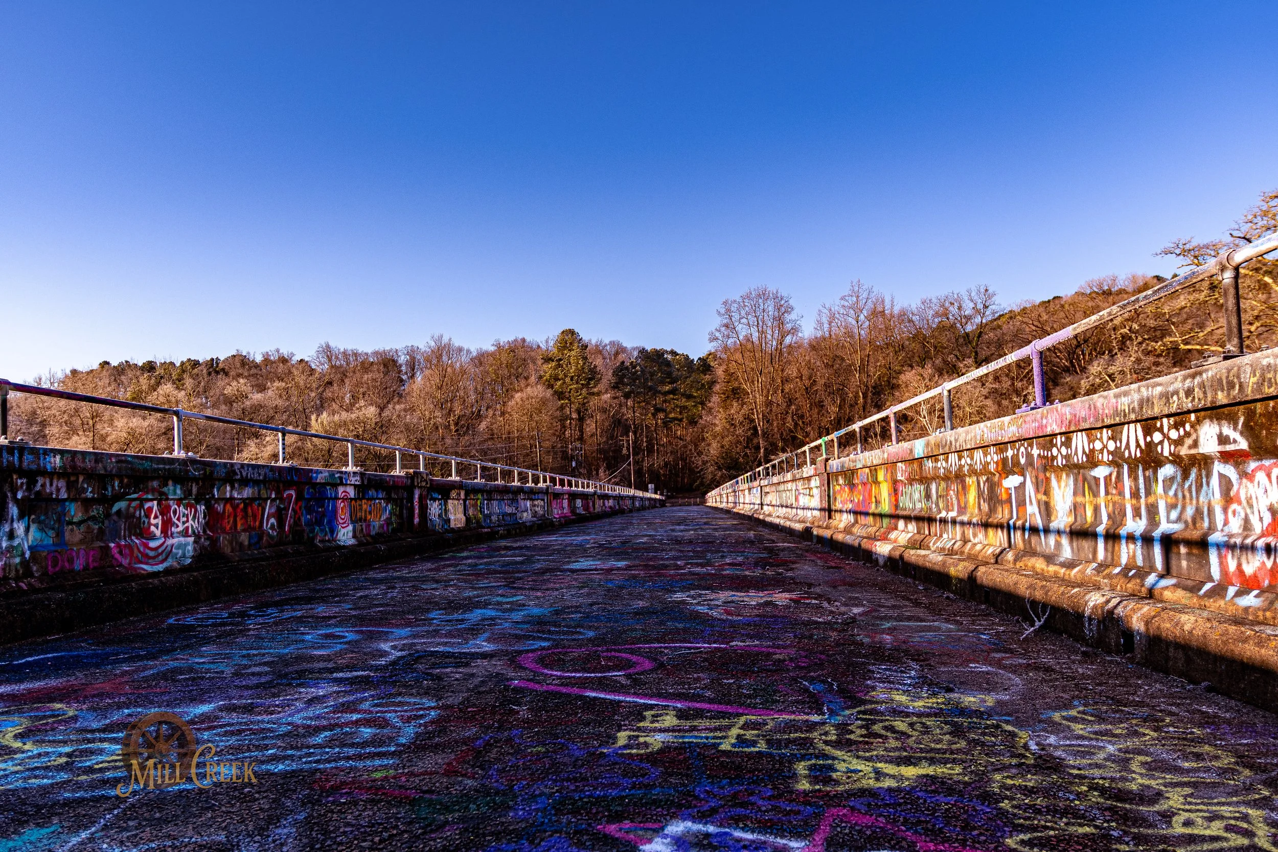 A graffiti-covered bridge with colorful spray paint on the sides and ground, surrounded by leafless trees and a clear blue sky.