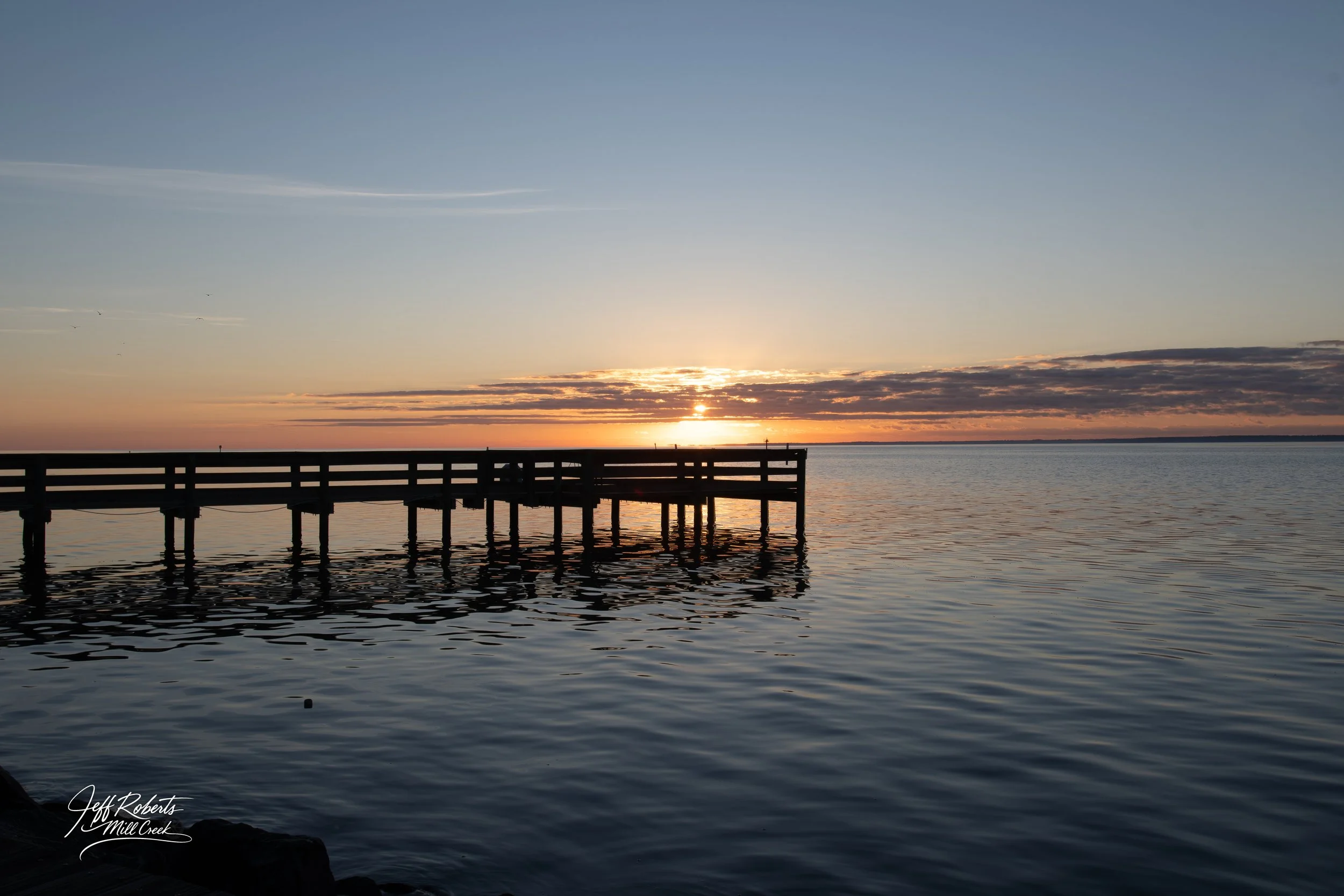 Sunset over a body of water viewed from a pier extending into the water, with colorful sky and clouds.