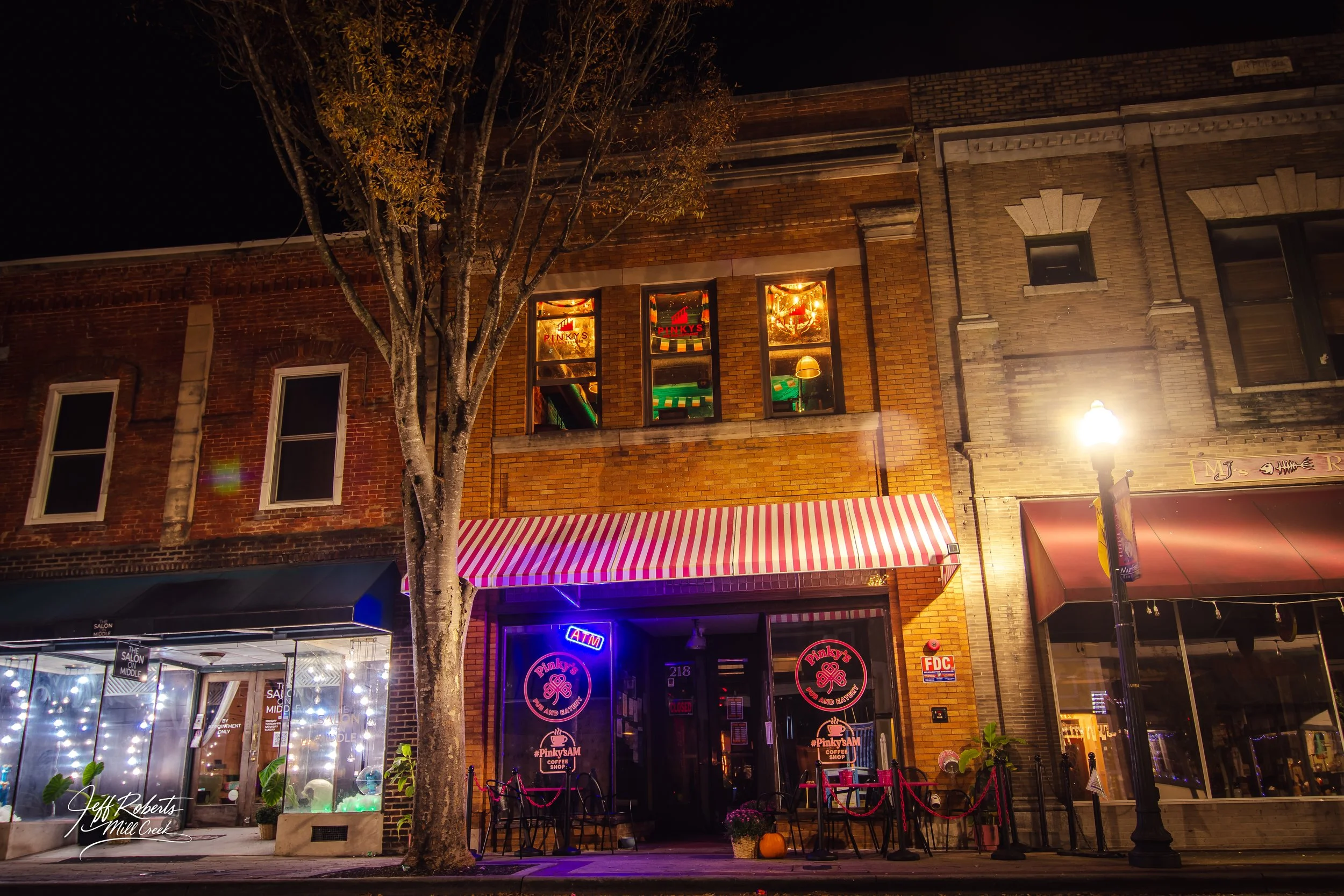 Night view of a brick storefront with striped awning, neon signs, and decorated windows, in a downtown area with a streetlamp and tree in the foreground.