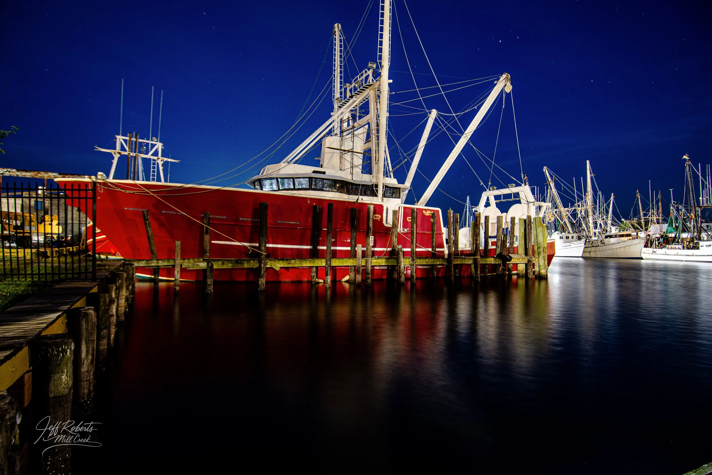 Nighttime view of wooden dock with red fishing boat and several sailboats docked in a marina under a starry sky