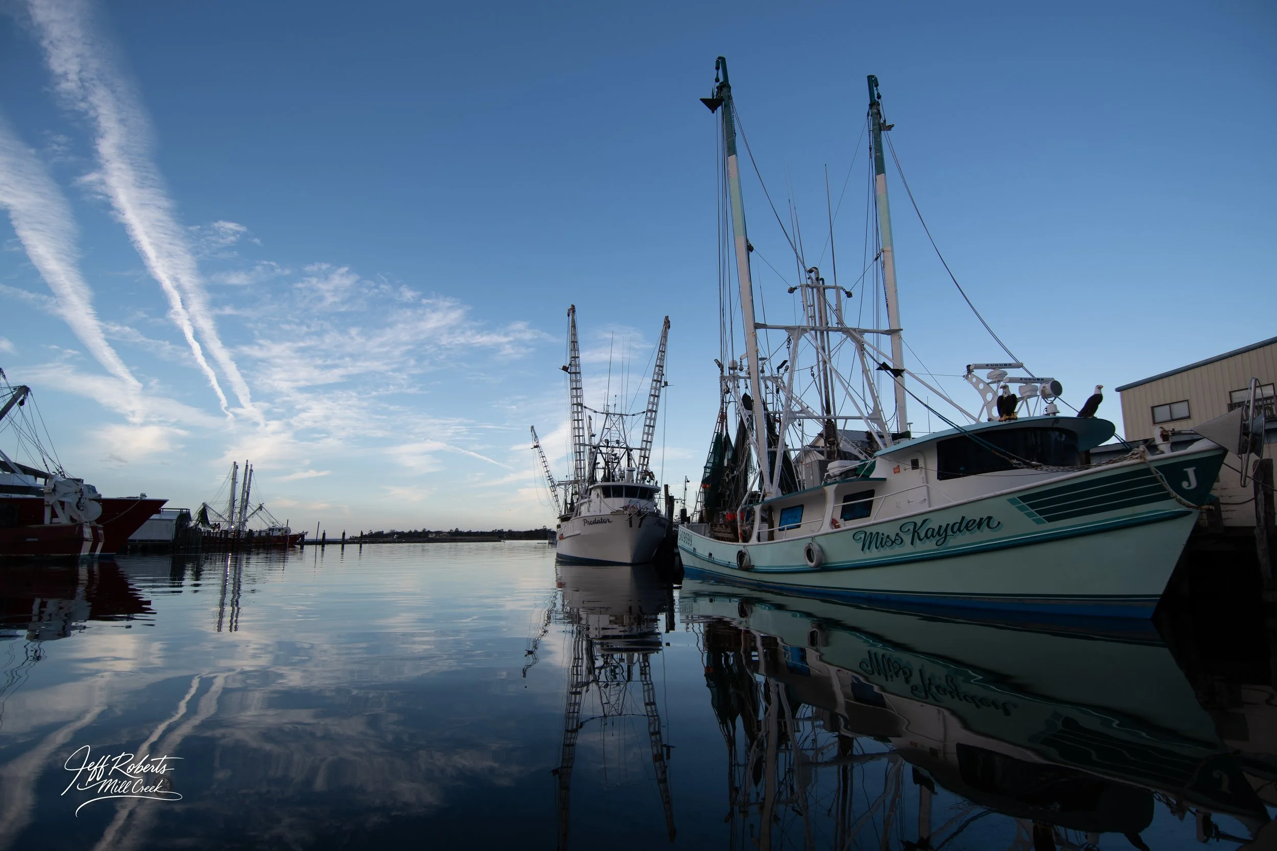 Boats docked at a marina on calm water with reflections, clear blue sky with wispy clouds, and a distant horizon.