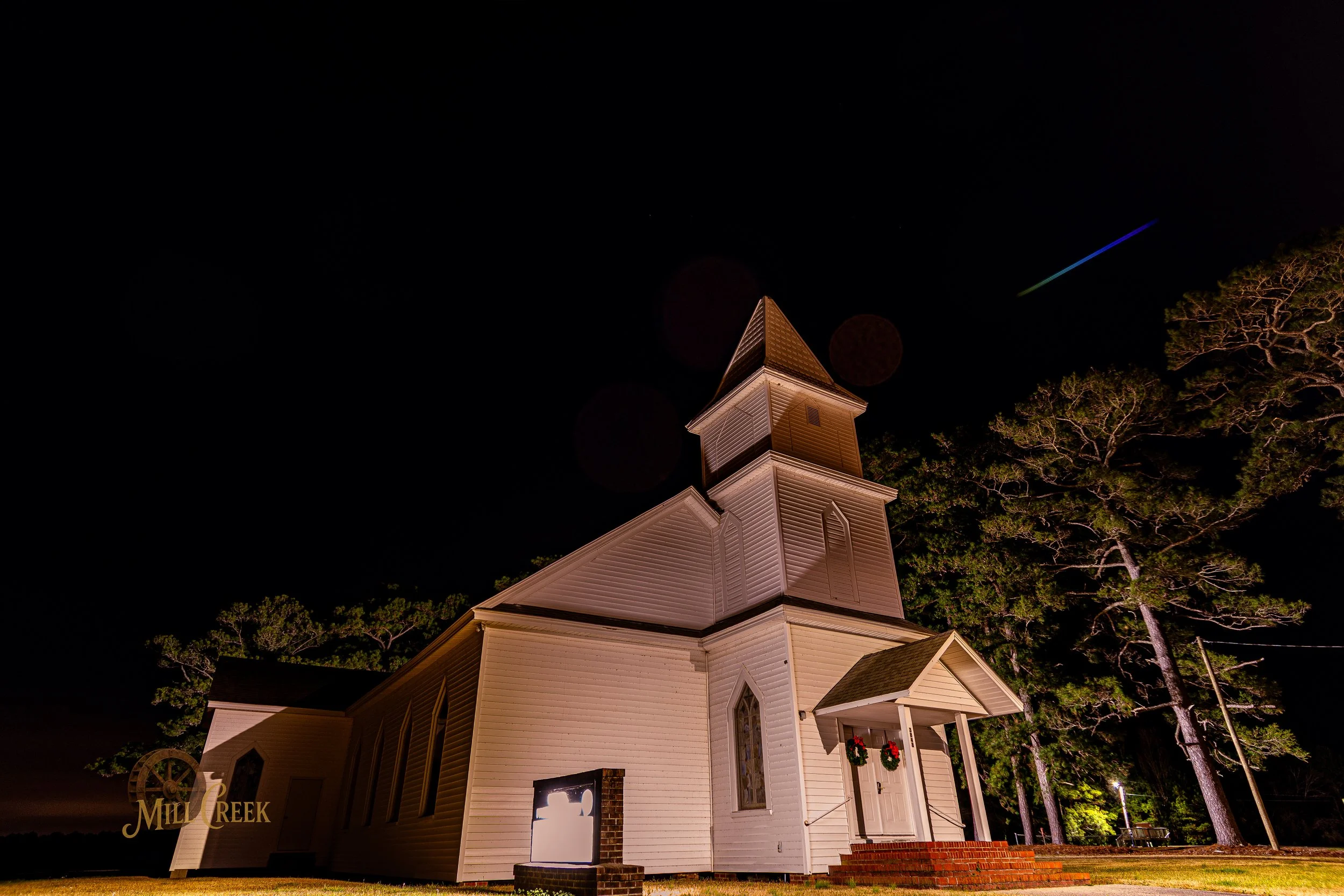 Nighttime exterior view of a white wooden church with a steeple, decorated with Christmas wreaths, surrounded by trees, illuminated at night.
