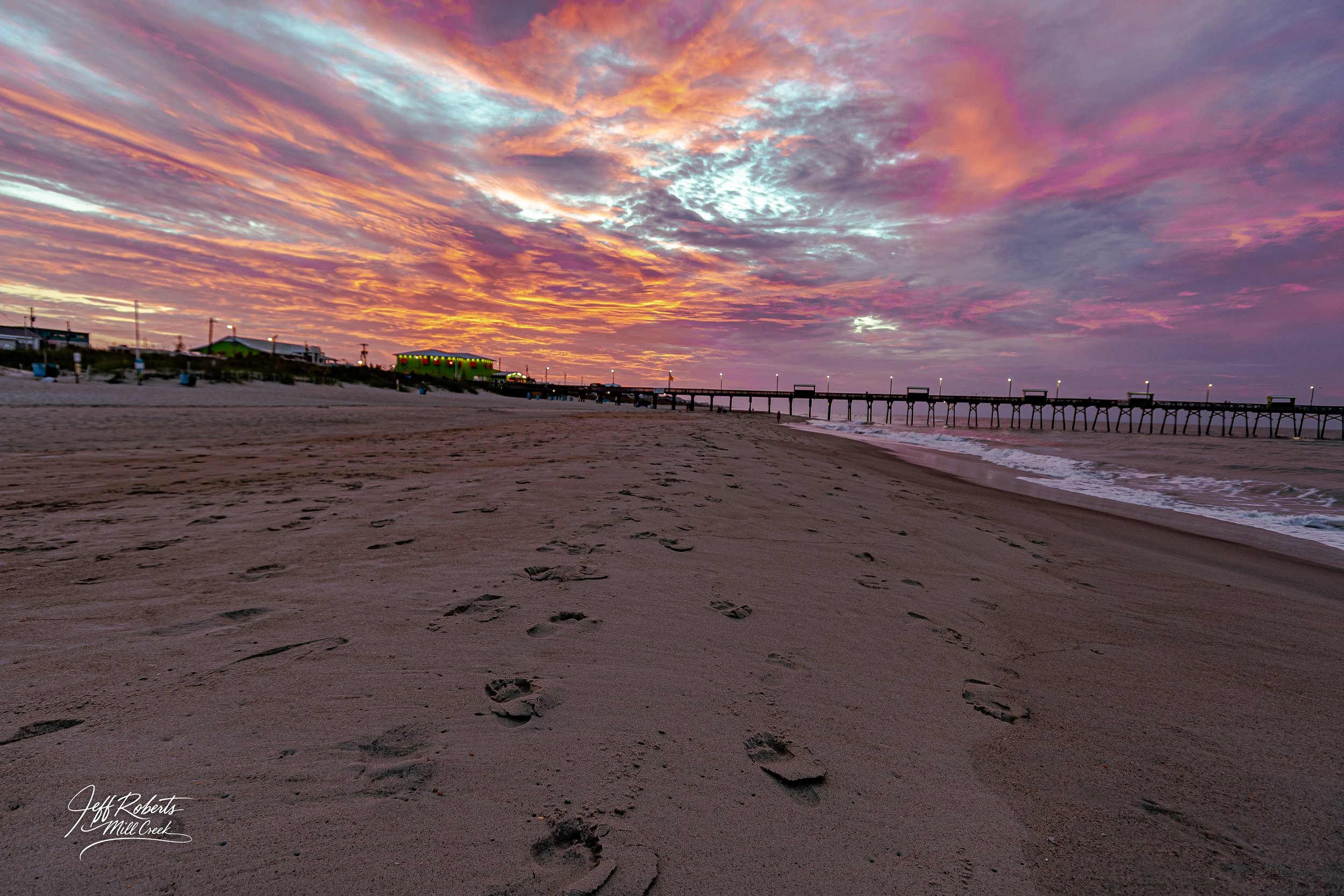 Footprints in the sand on a beach at sunset with a pier extending into the ocean and colorful sky with orange, pink, and purple clouds.