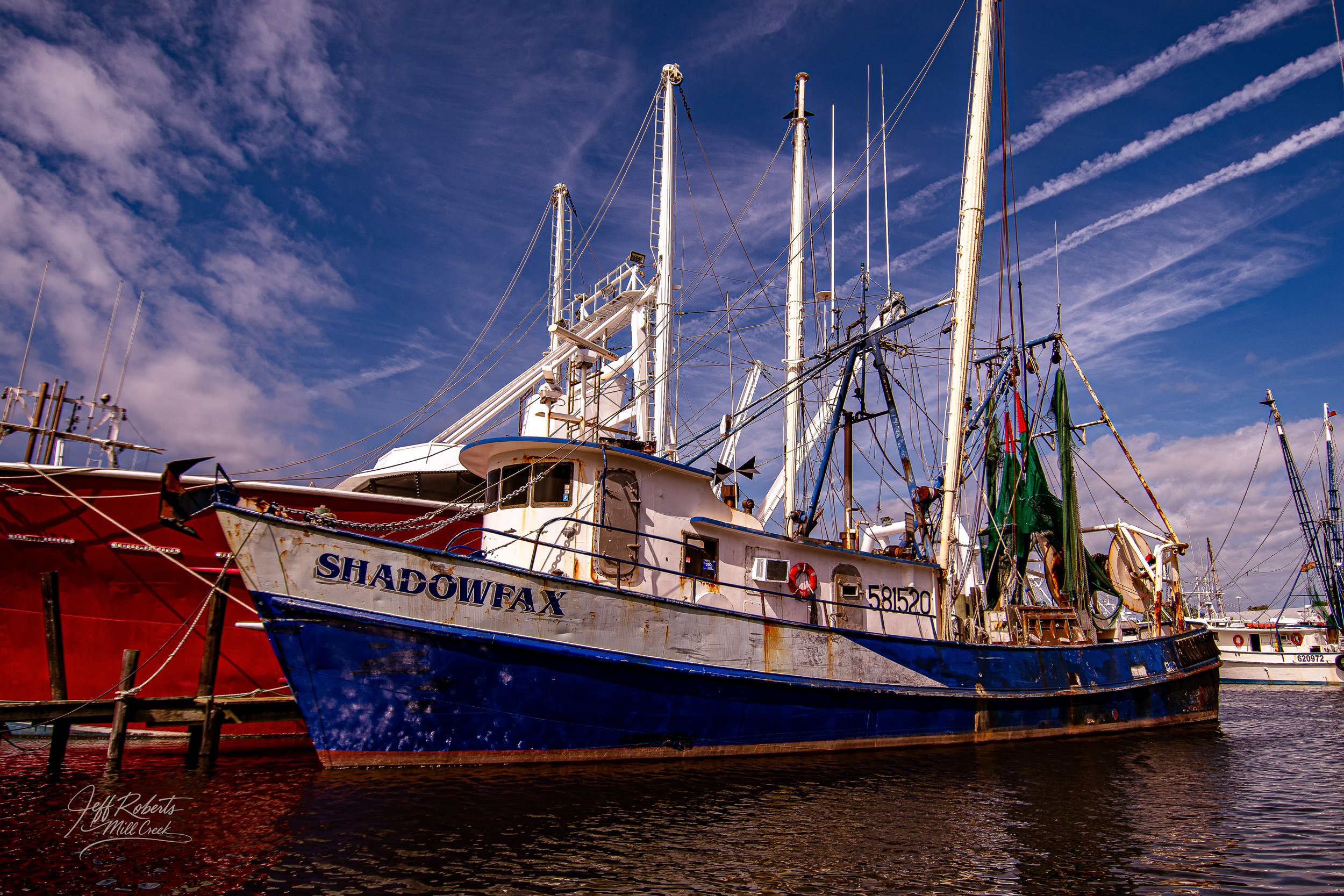 A fishing boat named 'Shadowfax' docked at a harbor, other boats and a blue sky with clouds in the background.