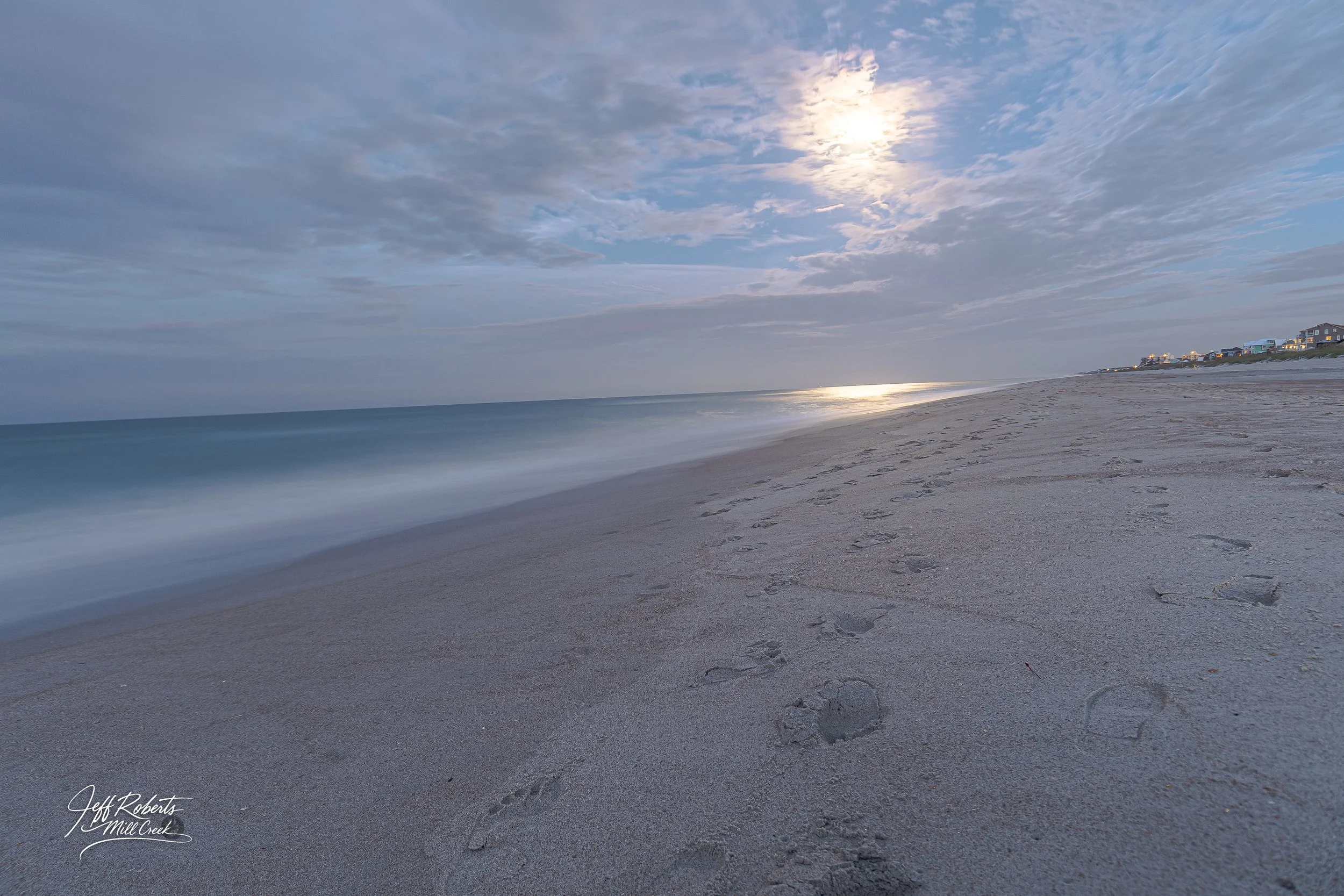 Empty sandy beach with footprints, ocean waves, cloudy sky, and houses in the distance at dusk.