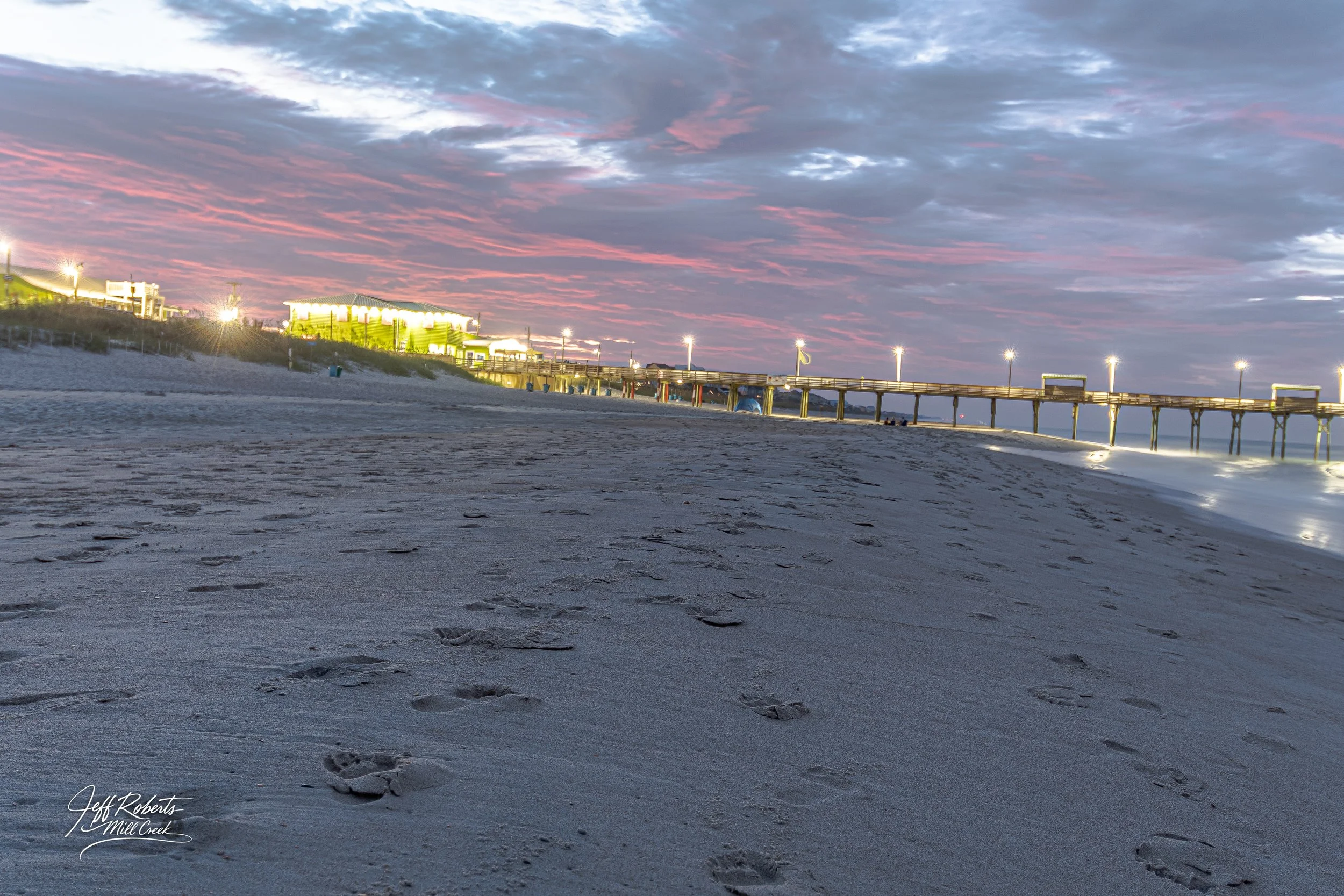 Empty sandy beach at sunset with footprints, pier with lights, and colorful sky with pink and purple clouds.