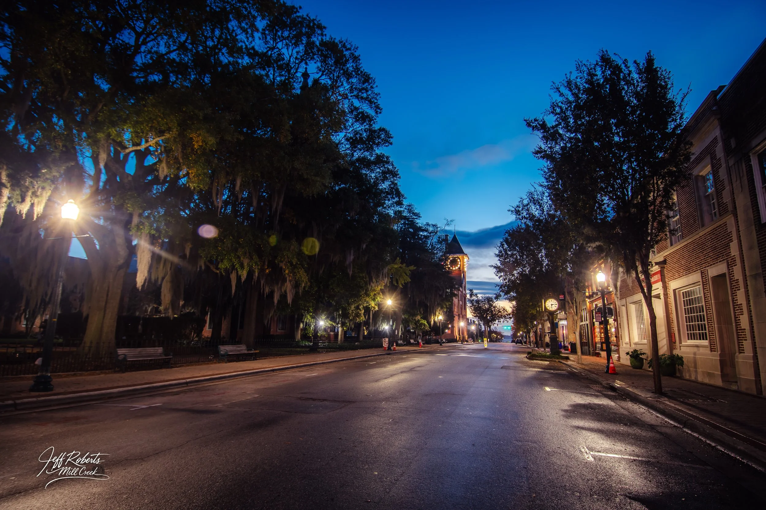 A quiet city street at dawn with streetlights on, trees on either side, and a clock tower in the distance under a blue sky.