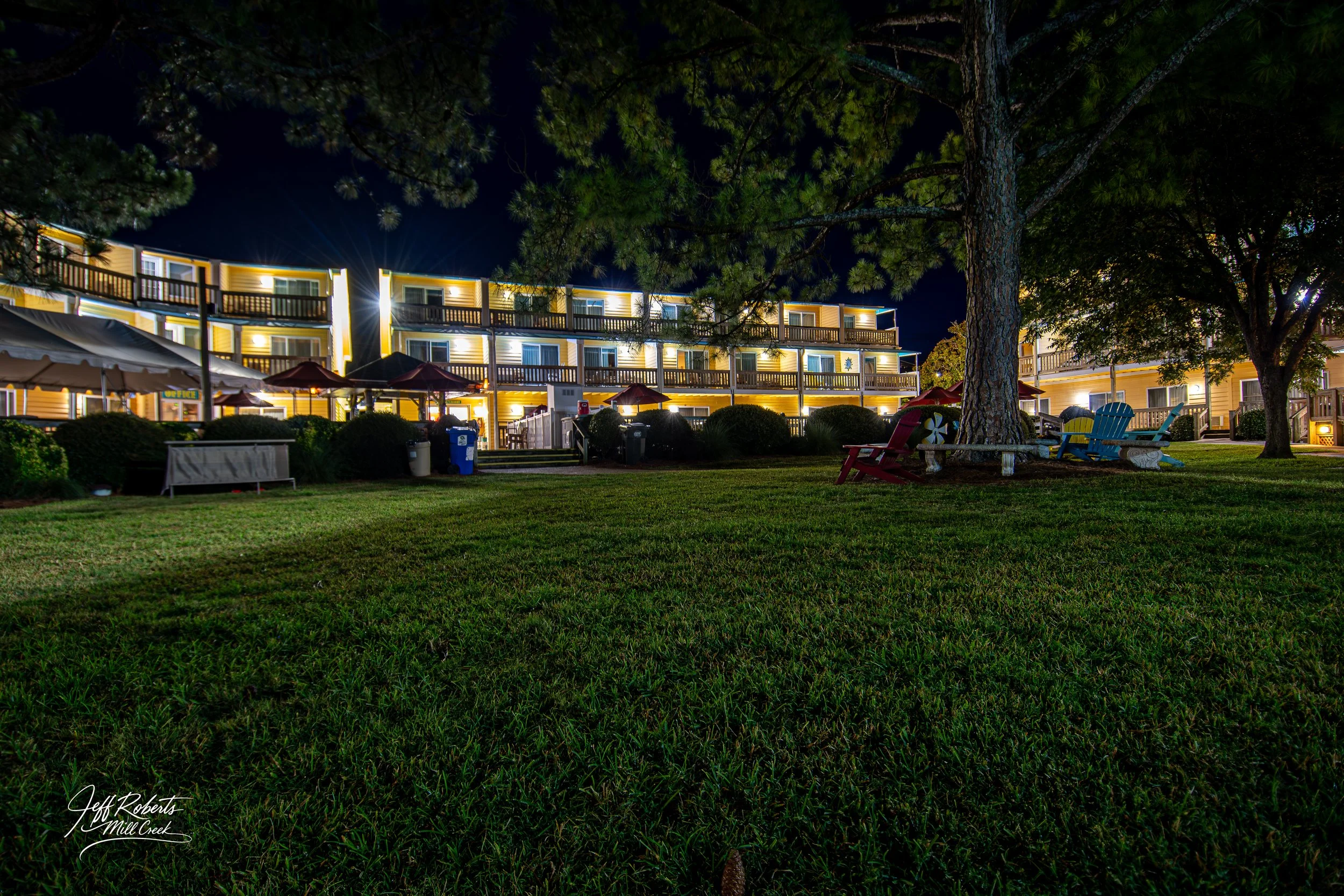 Night view of a motel with illuminated yellow two-story building, outdoor seating, and large trees on a well-kept lawn.
