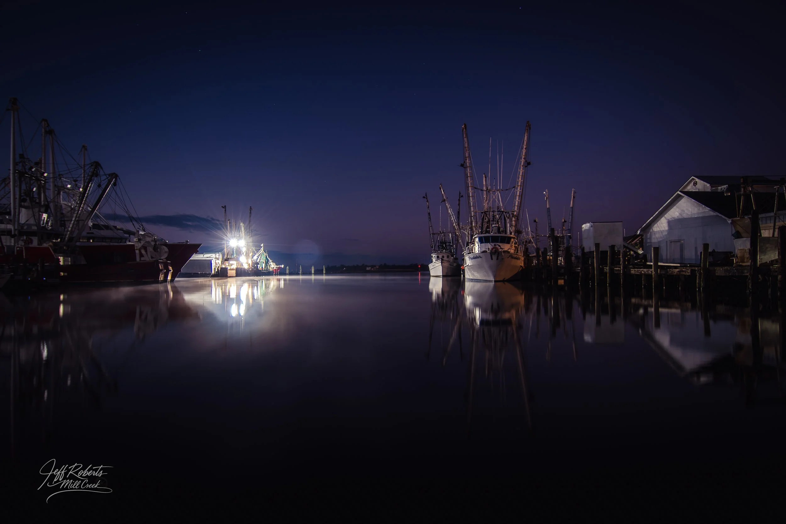 Nighttime view of boats docked at a harbor, calm water reflecting the boats and lights, clear sky with a few stars, industrial and residential buildings along the dock.