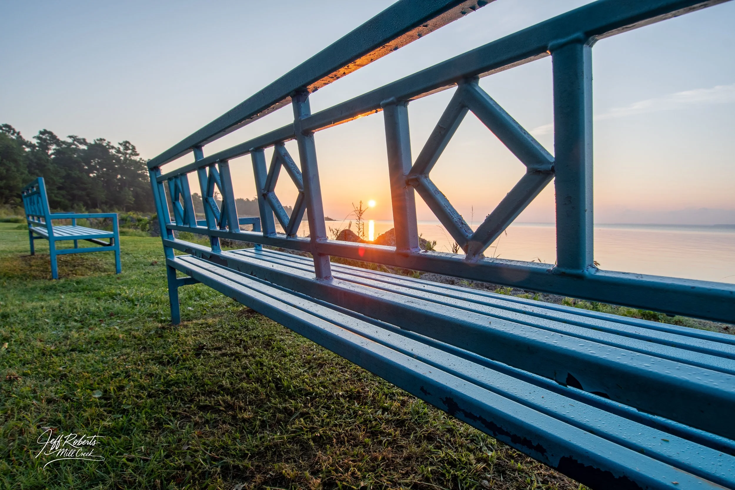 Blue benches facing a lake at sunset, with the sun near the horizon, trees in the background, and grass in the foreground.