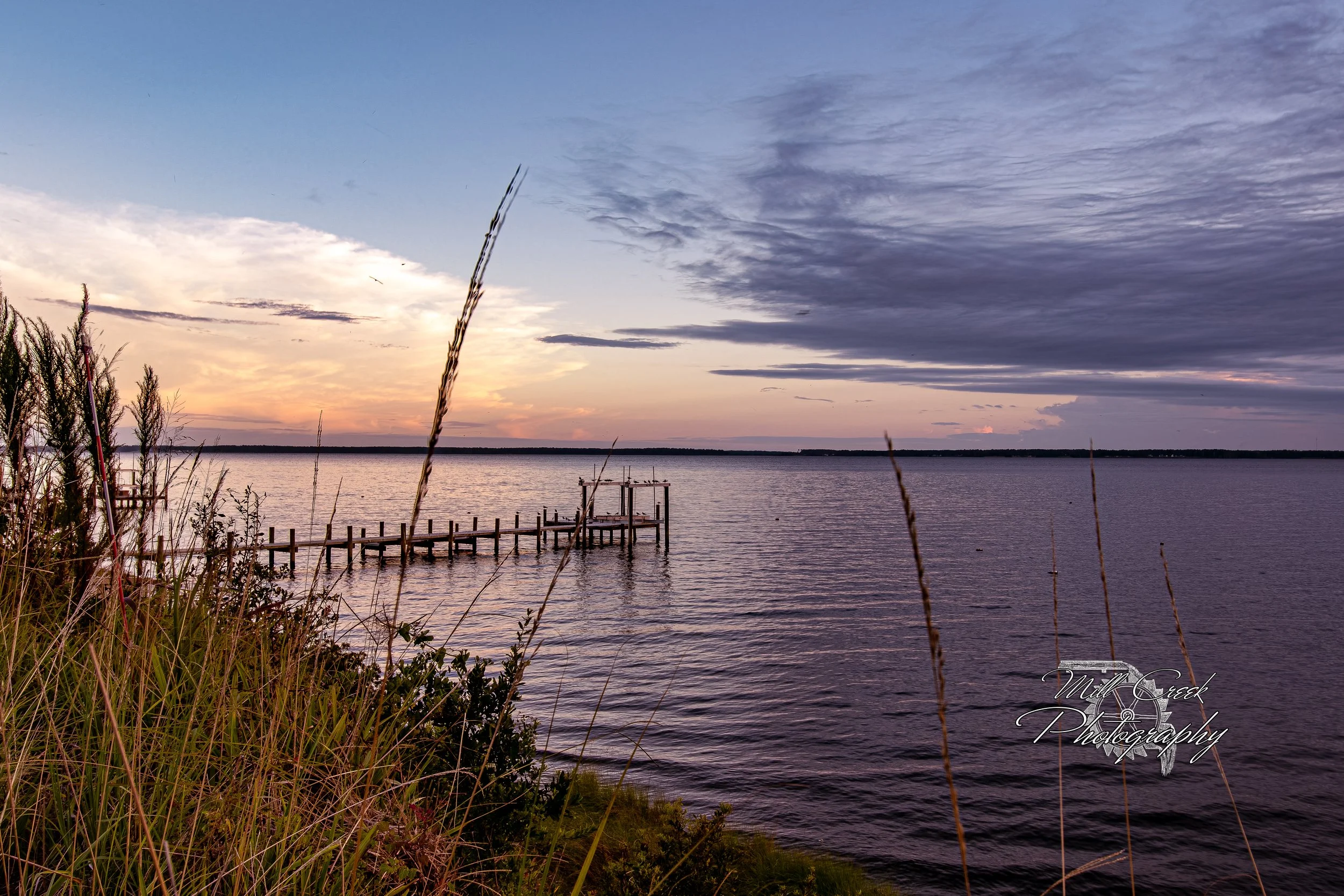 "Riverside Stillness"
Pastel evening light melts into calm water as a weathered dock reaches toward the horizon. Tall coastal grasses frame the scene while distant clouds glow with the last warmth of day.