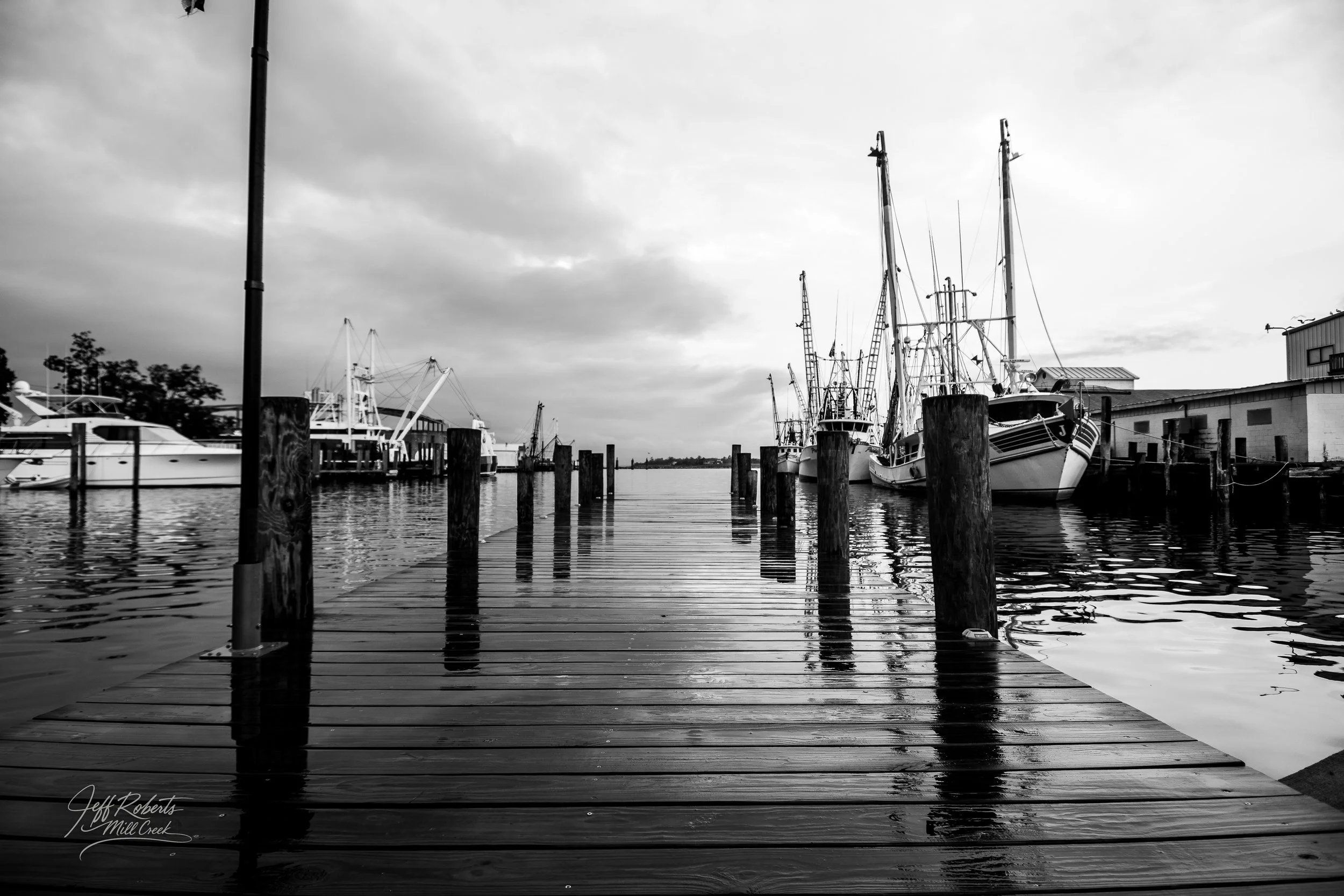 Black and white photo of a wooden dock extending into a harbor with boats and yachts moored on both sides, cloudy sky above.