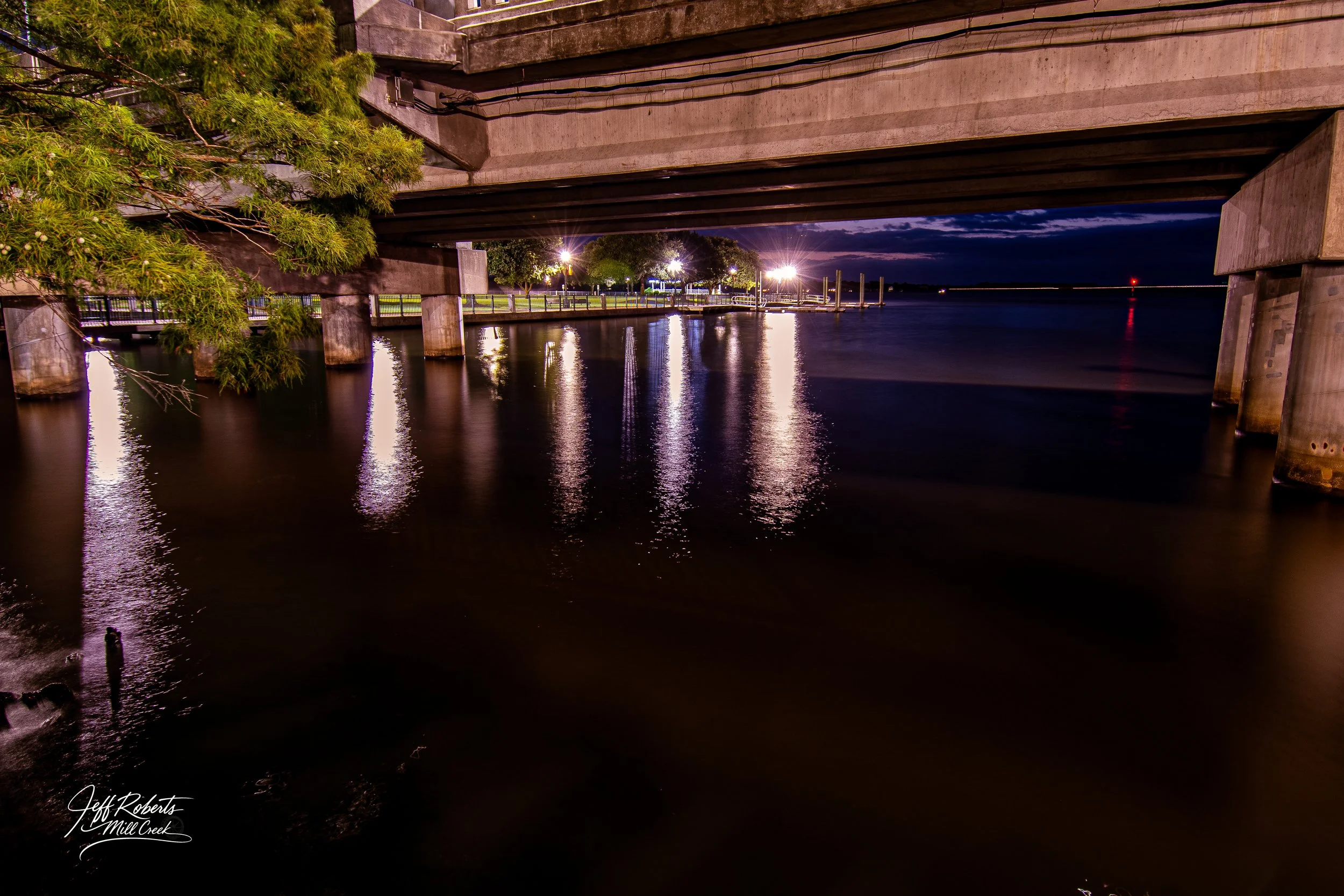 Night view underneath a bridge over a calm creek, with streetlights and trees along the water's edge, and a dark sky in the background.