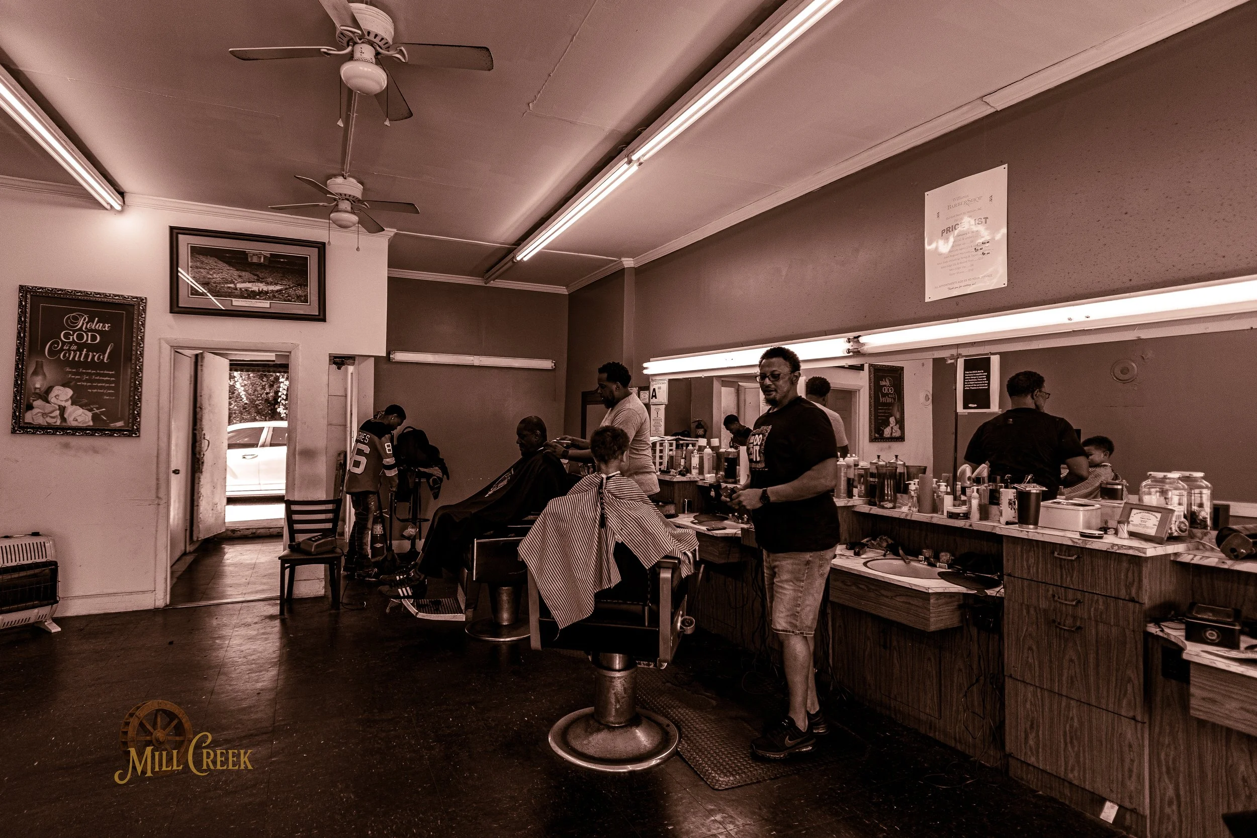 Inside a barbershop with clients getting haircuts. Barbers are working at the counter with hair products and tools. A man is standing near a client in a barber chair, and another client is seated. There are ceiling fans and framed pictures on the wal