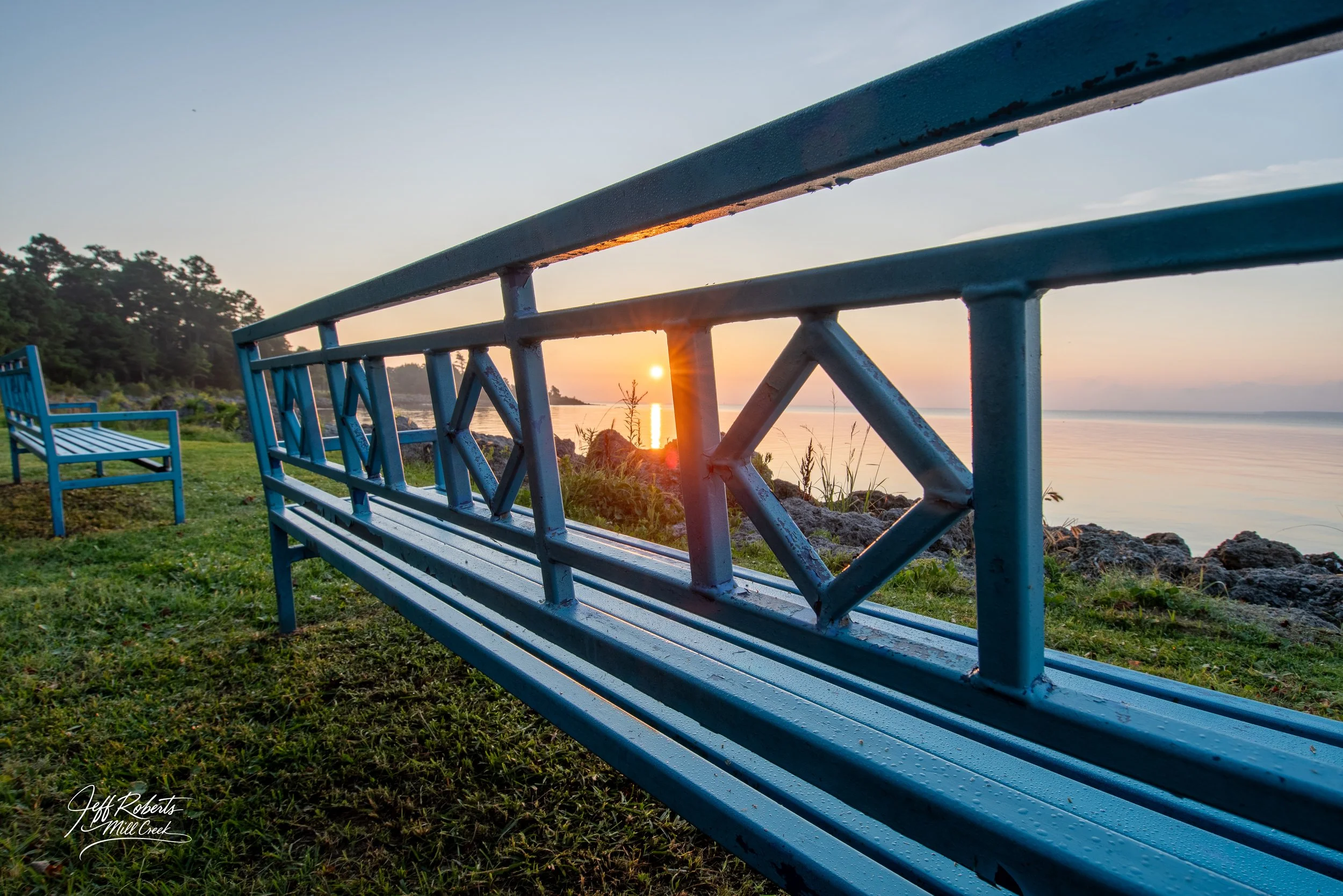 Blue benches on grassy area near rocky shoreline with sunset over calm water and a distant tree-lined horizon.