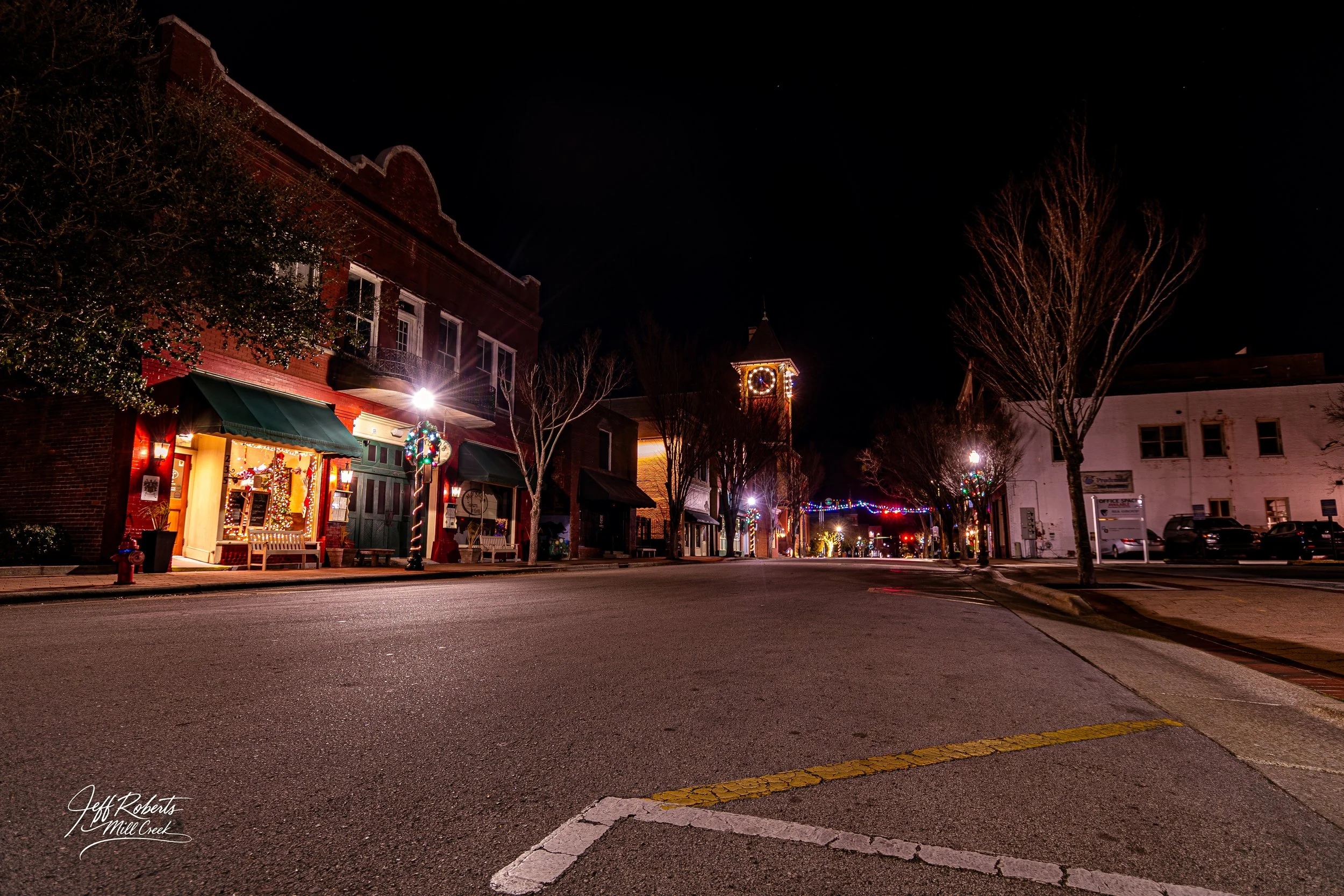 Empty street in a small town at night with holiday lights and decorations, including a decorated lamppost, with a clock tower in the background.