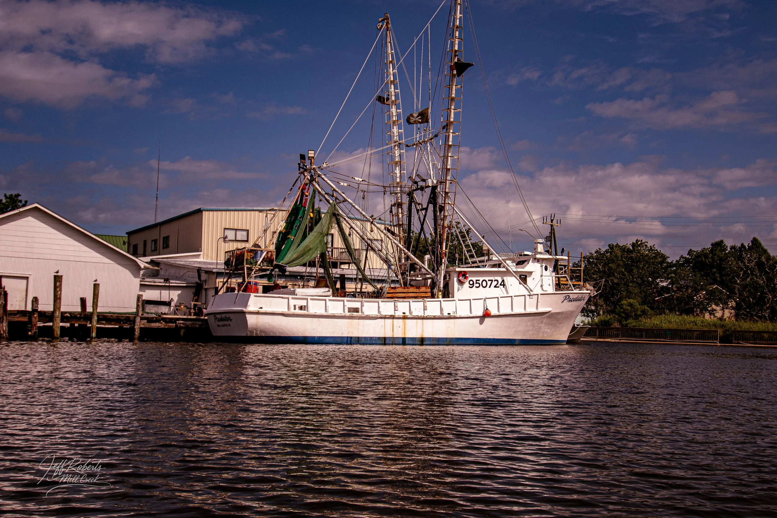 A white fishing boat named Predator docked at a pier with buildings and trees in the background, under a partly cloudy sky.