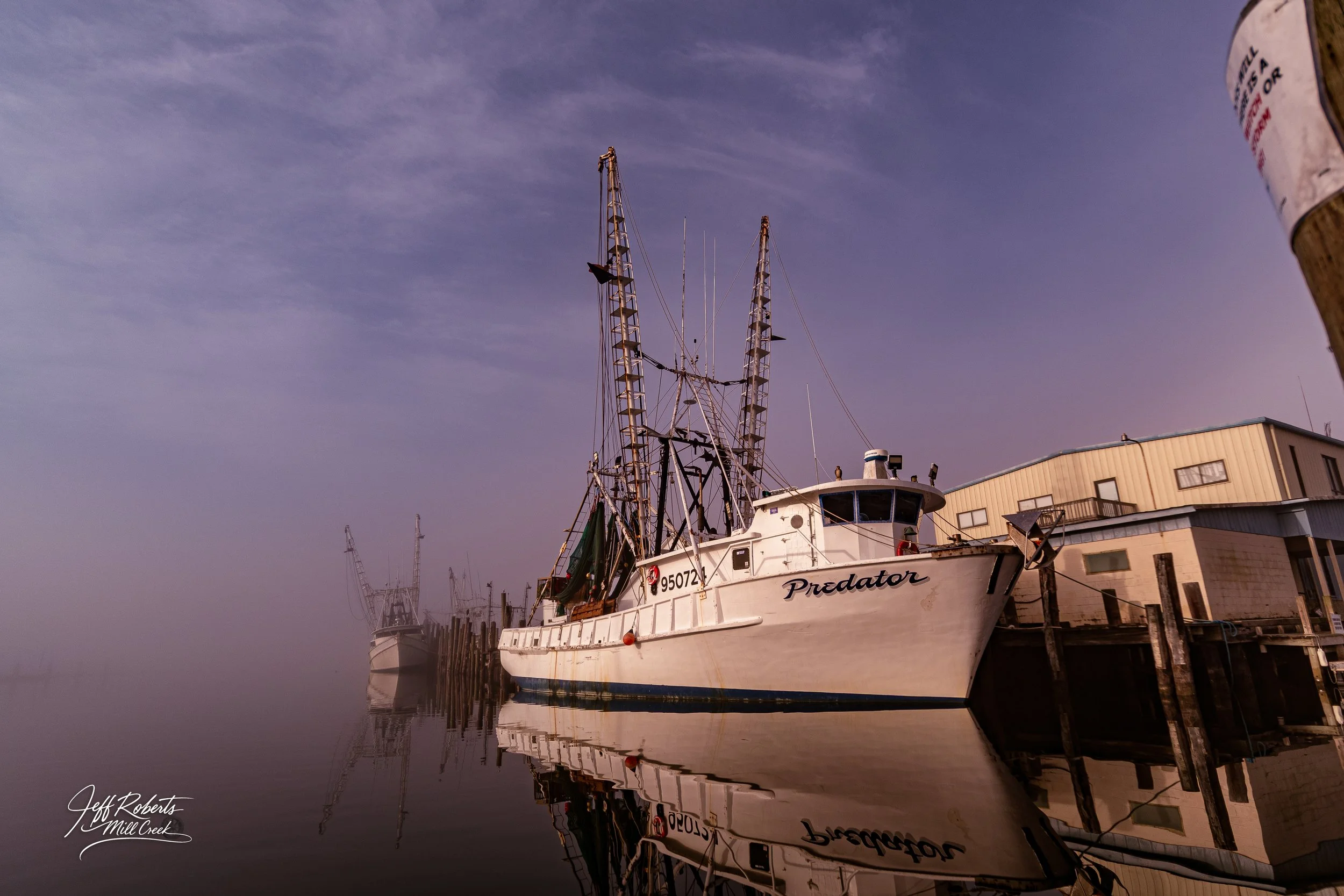A white fishing boat named Predator docked at a pier with calm water and an overcast sky.