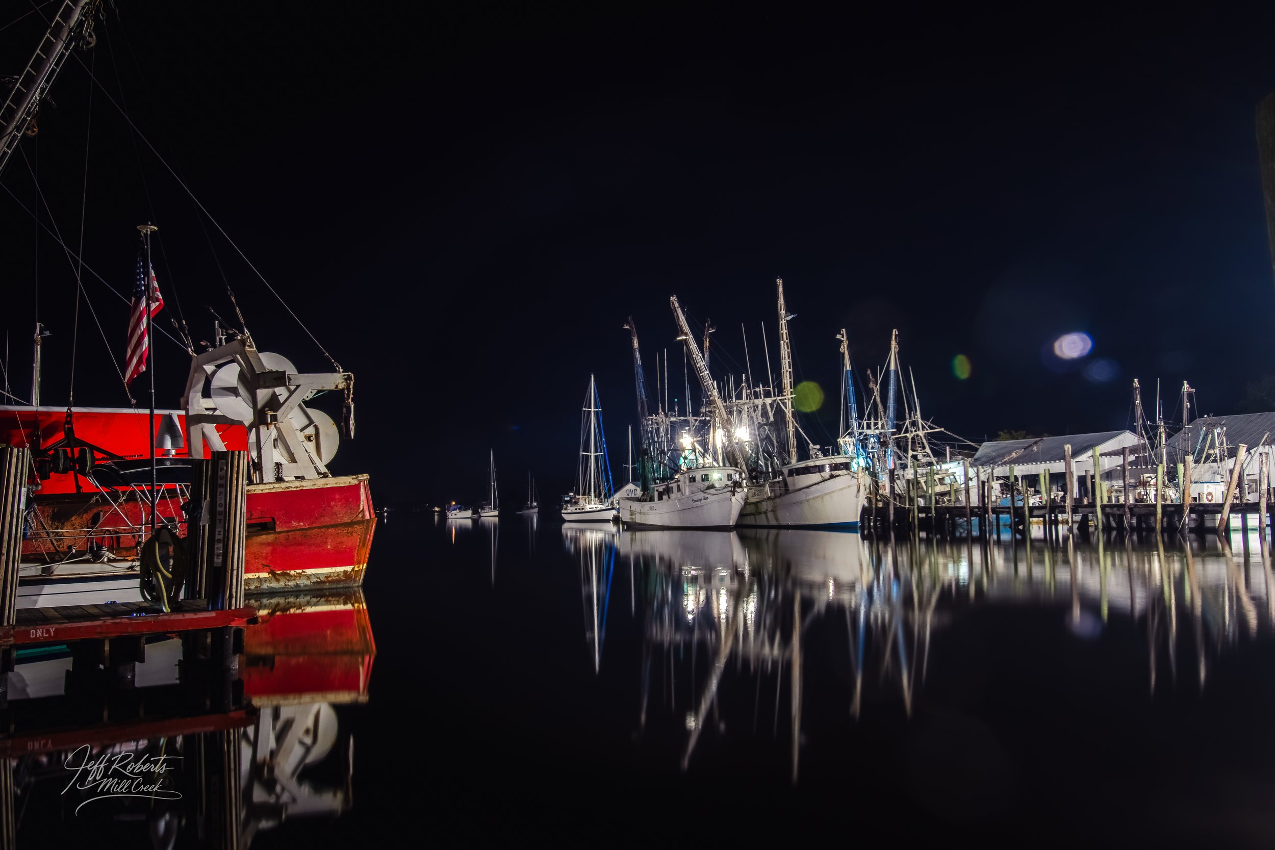 Nighttime scene of docked boats and yachts at a marina, with reflections on the calm water, illuminated by boat lights.