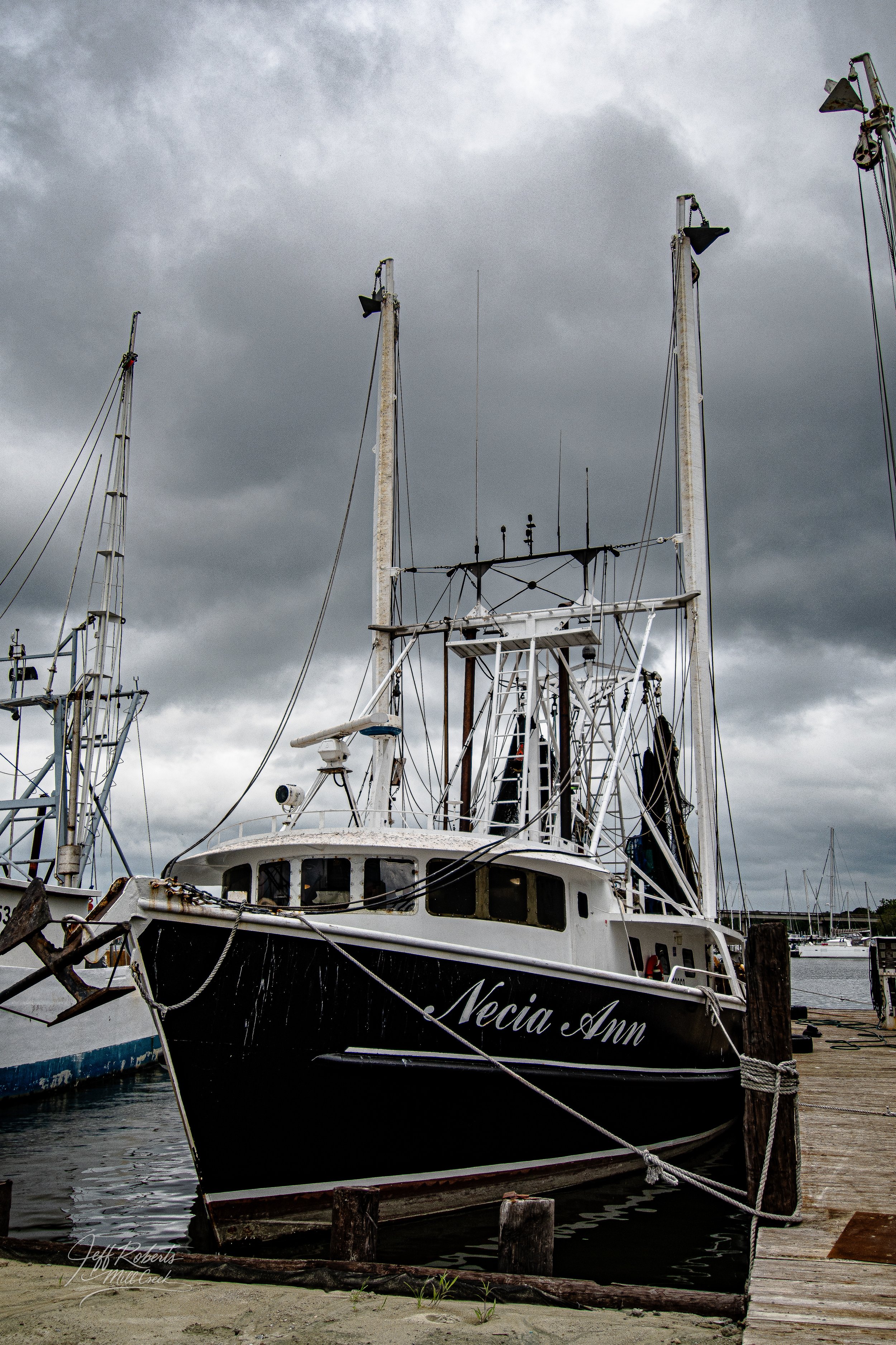 A boat named Necia Ann docked at a marina under dark, cloudy skies, alongside other boats.